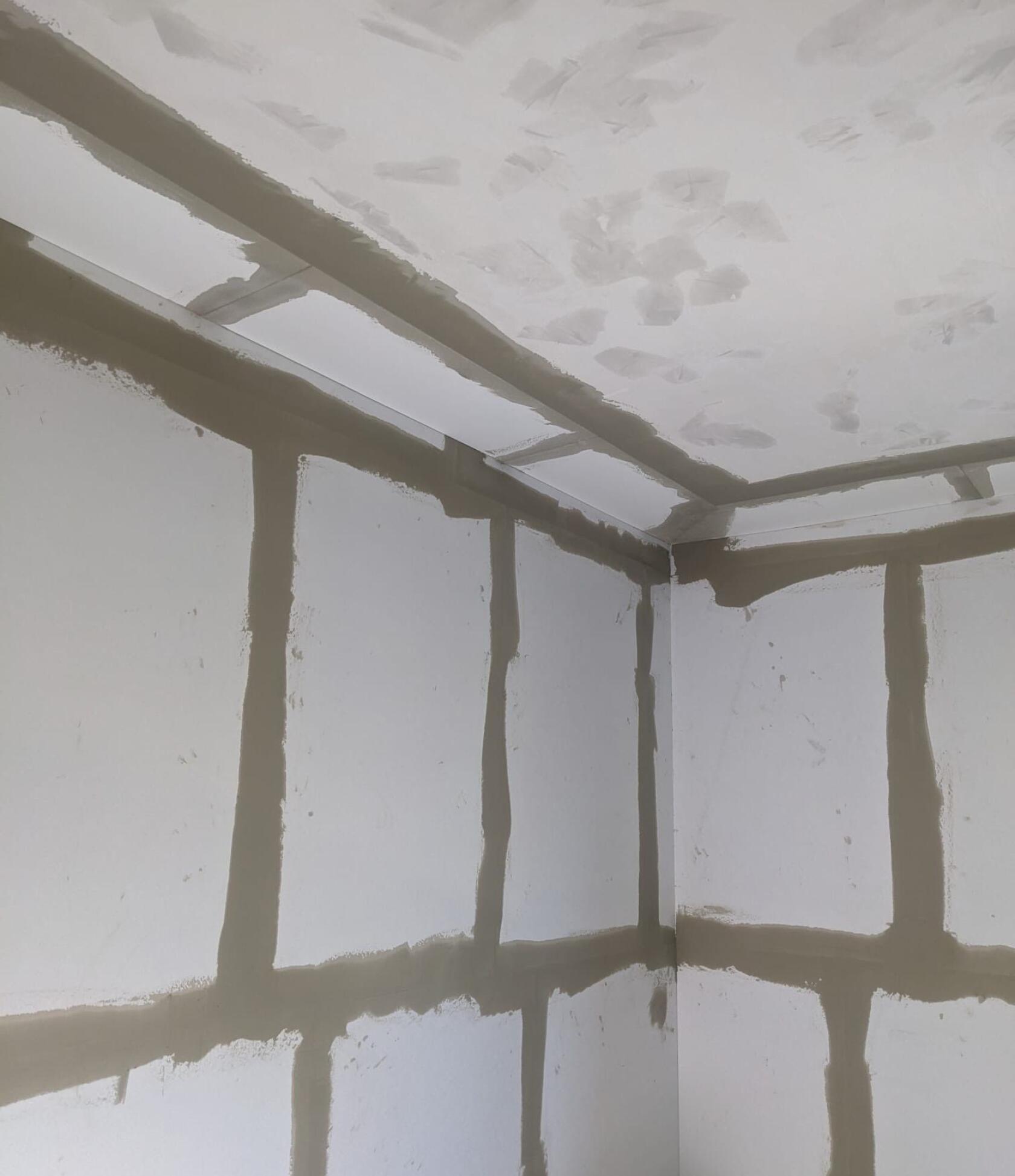 Gipskartonplatten in einer Raumecke – Fugen und Ecken sind mit beigefarbener Spachtelmasse verspachtelt – weiße Wand- und Deckenplatten mit sichtbaren vertikalen und horizontalen Nahtstreifen und Deckenflicken.