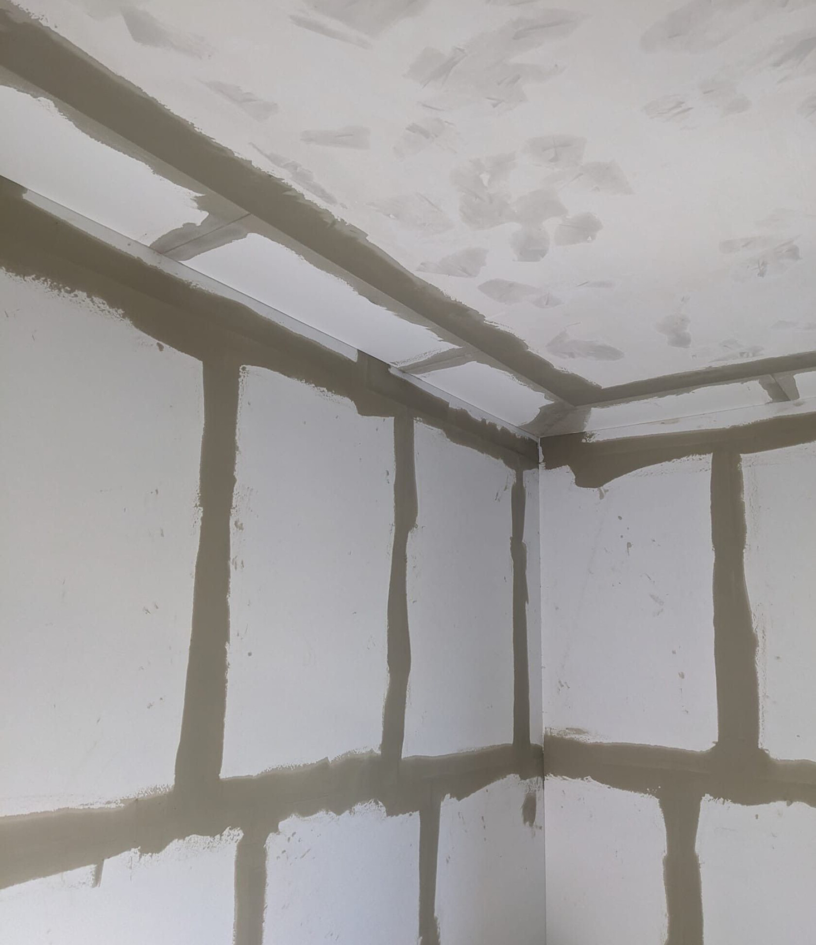 Gipskartonplatten in einer Raumecke – Fugen und Ecken sind mit beigefarbener Spachtelmasse verspachtelt – weiße Wand- und Deckenplatten mit sichtbaren vertikalen und horizontalen Nahtstreifen und Deckenflicken.