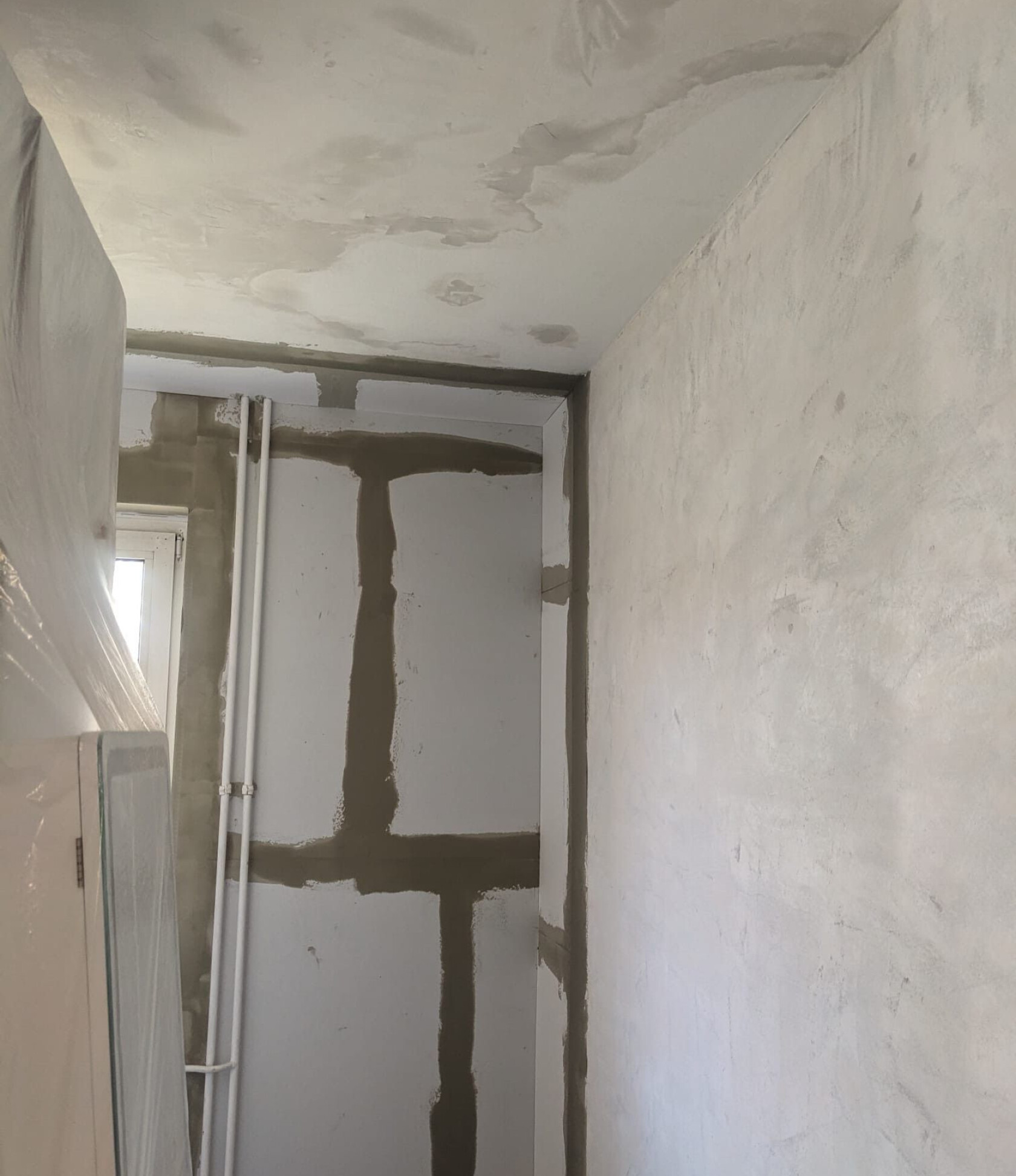 Raumecke – gespachtelte Fugen bilden an der linken Wand ein dunkles Kreuzmuster, zwei senkrechte weiße Rohrleitungen, fleckige Decke mit Verfärbungen, rechts glatter Putz.