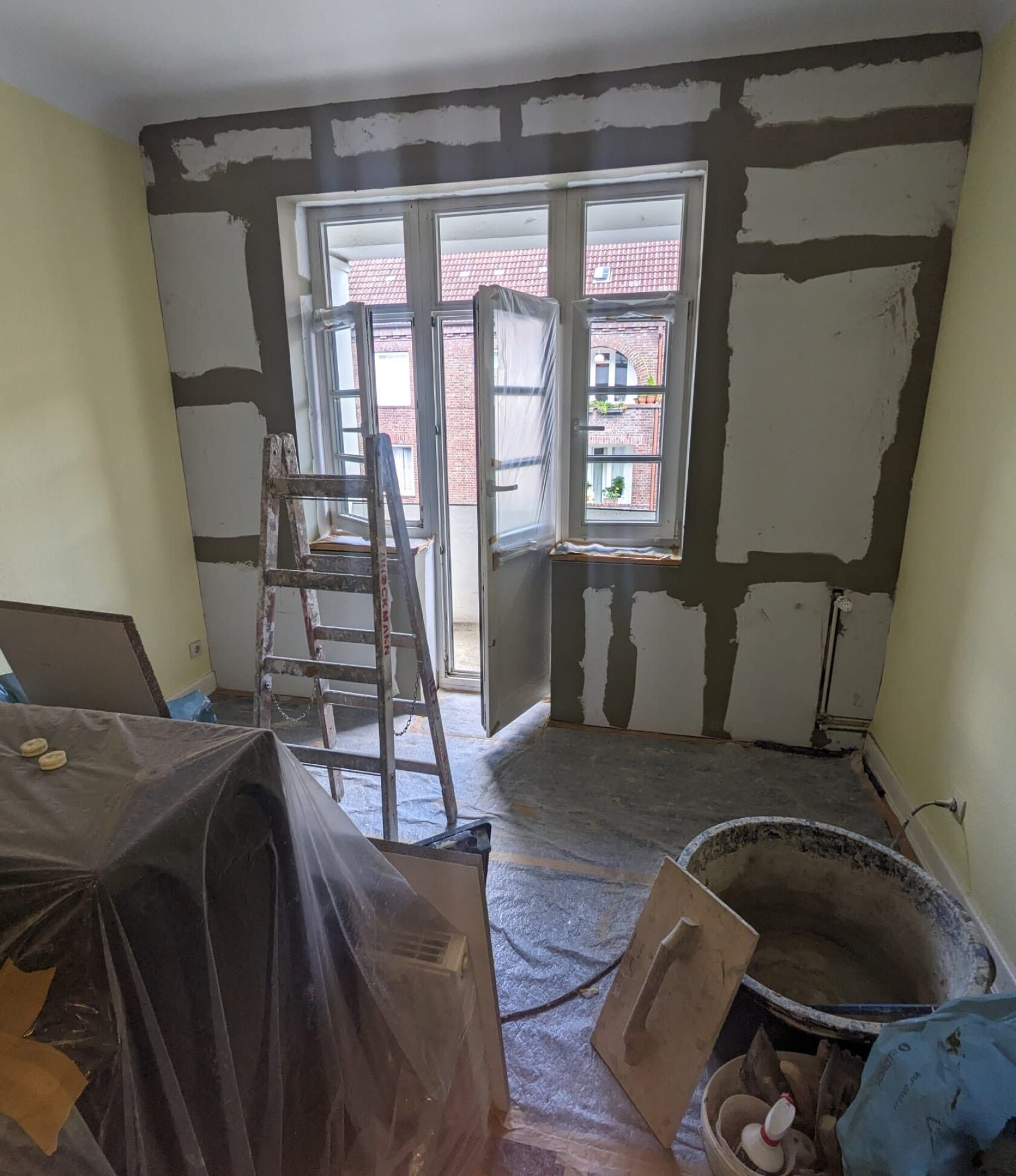 Aluminiumleiter steht vor einer geöffneten Balkontür; umgebende Wand mit Spachtelstellen, Boden und Möbel mit Folie abgedeckt, Eimer, Glättkelle und Bauwerkzeuge im Raum.