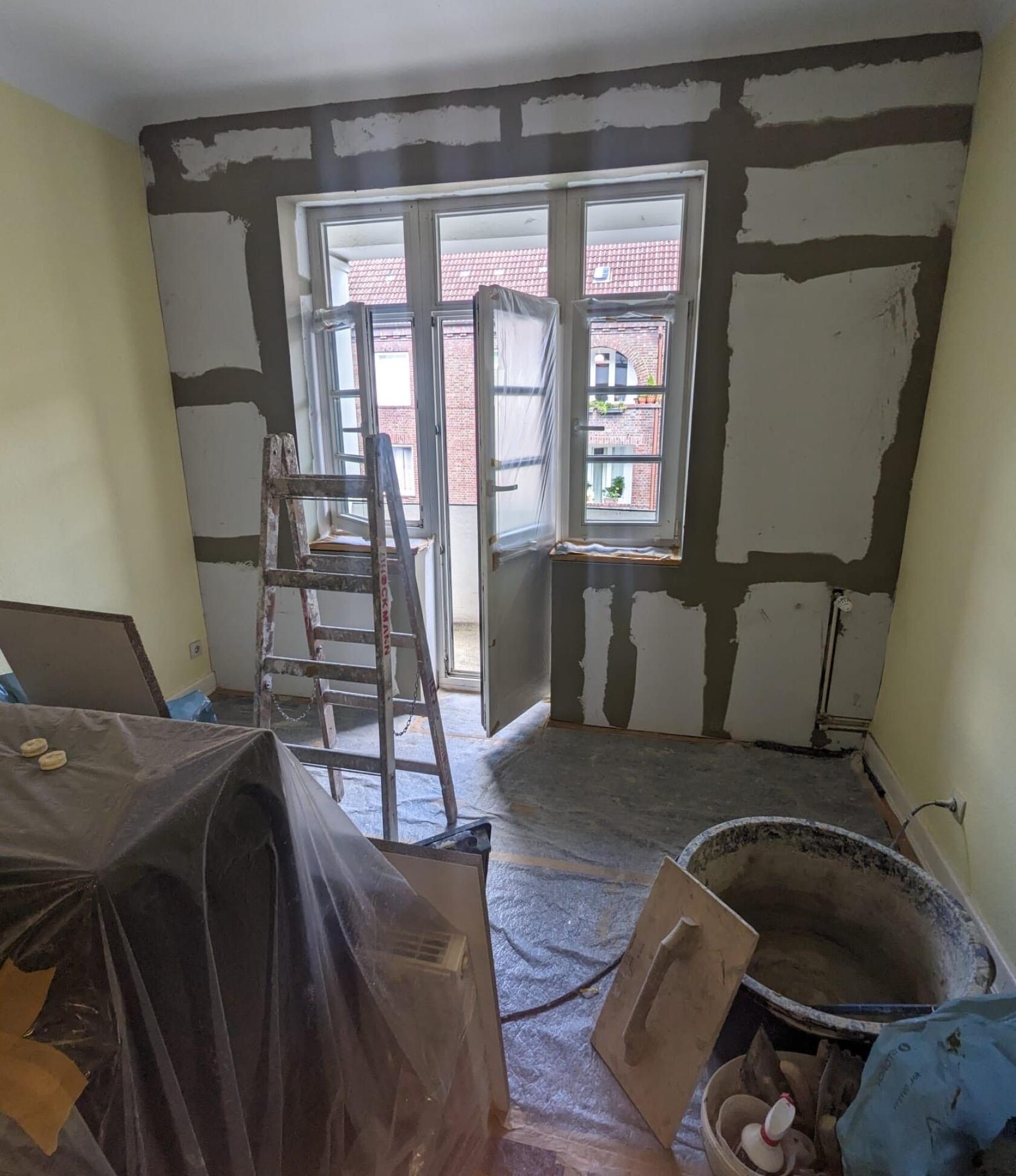 Aluminiumleiter steht vor einer geöffneten Balkontür; umgebende Wand mit Spachtelstellen, Boden und Möbel mit Folie abgedeckt, Eimer, Glättkelle und Bauwerkzeuge im Raum.