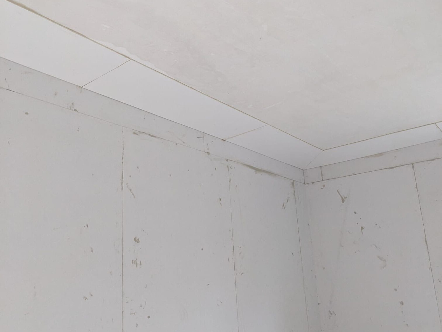 Ecke eines weißen Rohbauzimmers: Gipskartonwände und Deckenplatten treffen sich, sichtbare Nähte und Fugen, schmutzige Flecken und Putzspuren an den Wandflächen.
