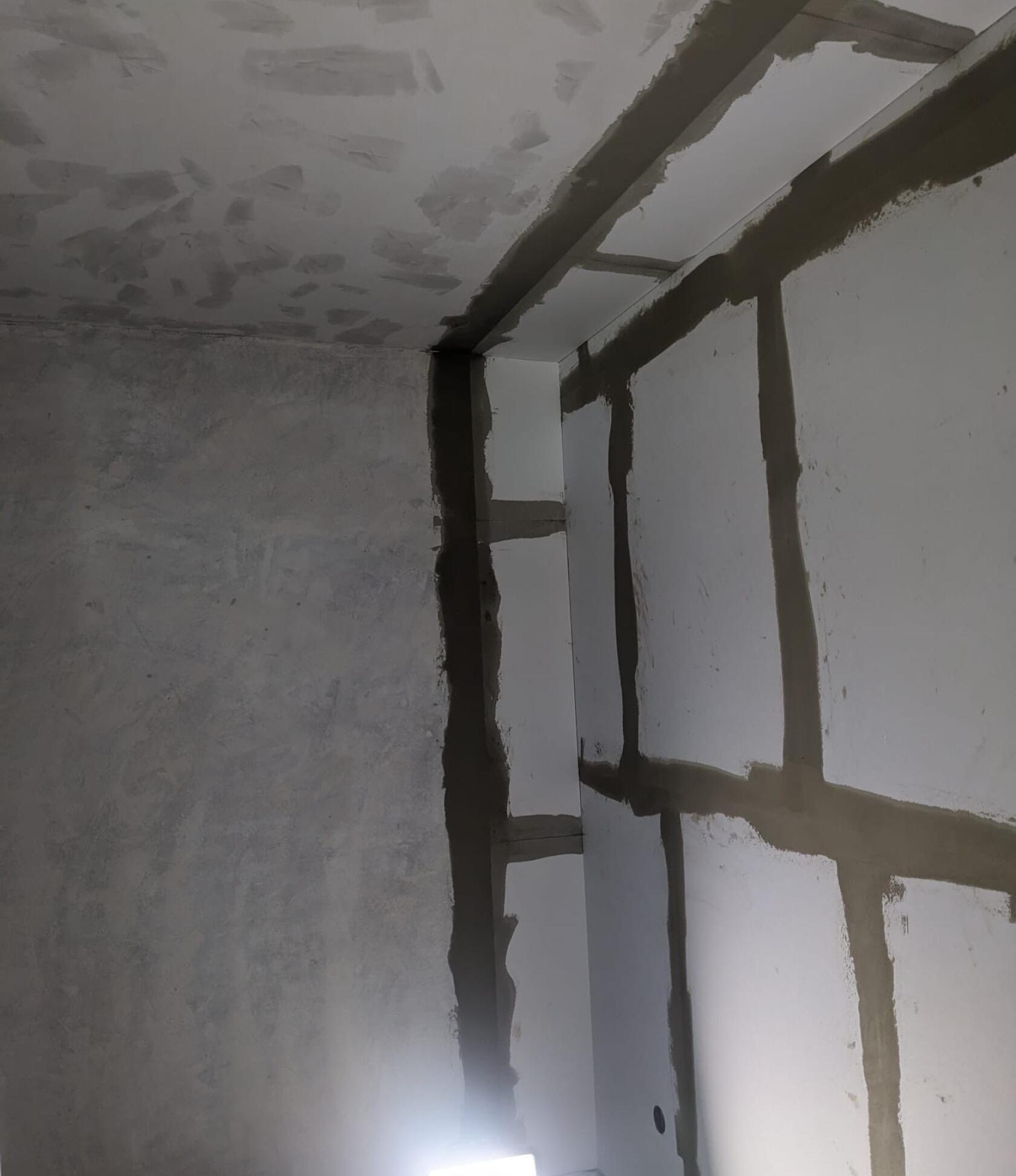 Ecke eines Innenraums — weiße Gipsplatten mit breiten, dunkelgrauen verspachtelten Fugen senkrecht und waagerecht; Decke mit unregelmäßigen Putzstellen; kleine Lichtquelle unten.