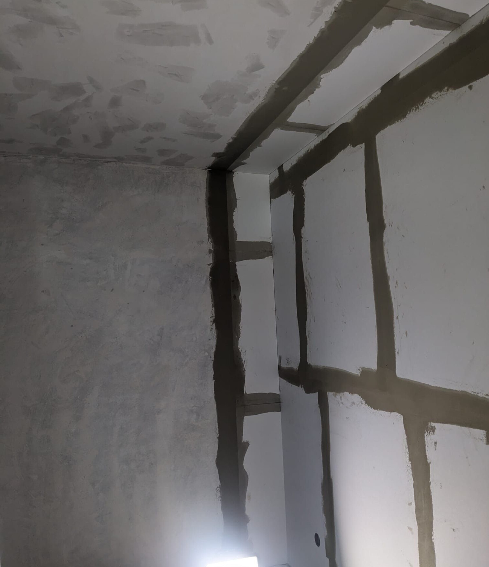 Ecke eines Innenraums — weiße Gipsplatten mit breiten, dunkelgrauen verspachtelten Fugen senkrecht und waagerecht; Decke mit unregelmäßigen Putzstellen; kleine Lichtquelle unten.