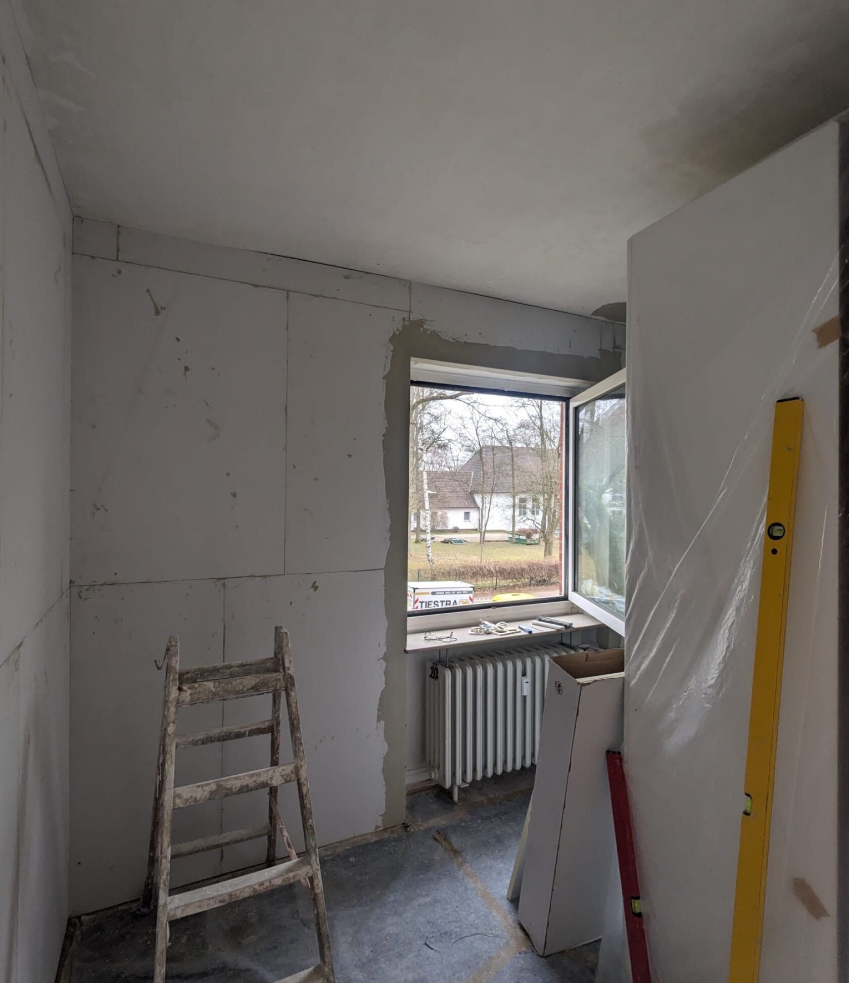 Alte Holzleiter steht auf Boden; in einem Renovierungsraum mit unvollständigen Gipskartonwänden, offenem Fenster über Heizkörper, Außensicht auf Haus und Schild 