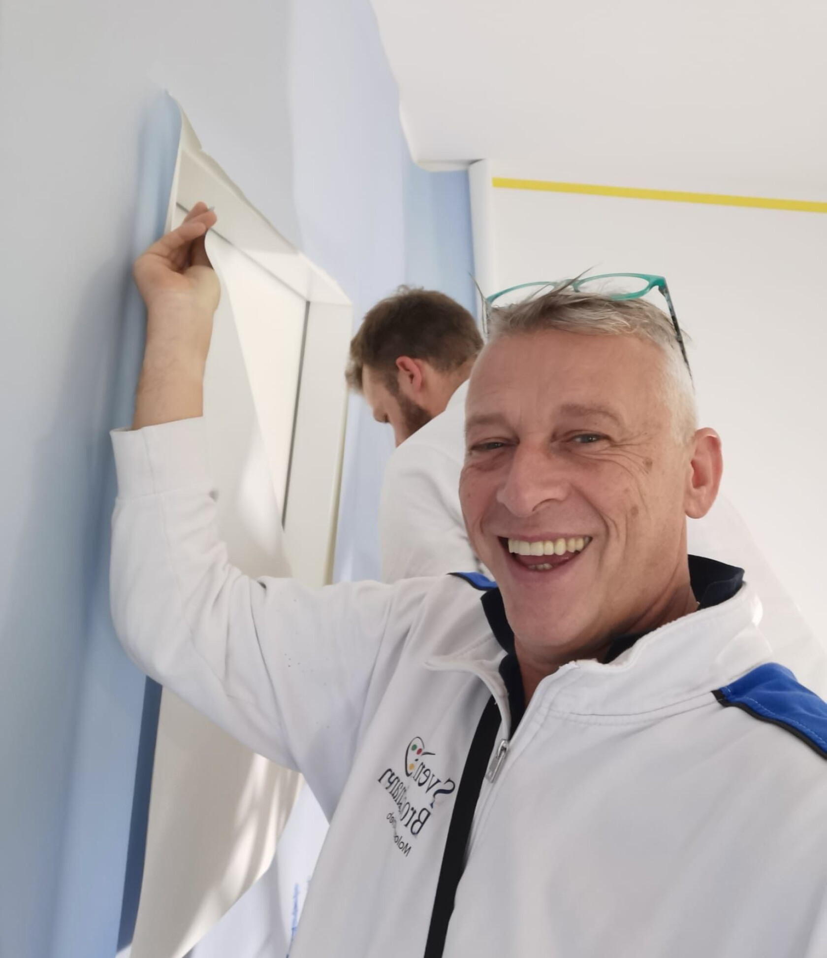 Vordergrund: Mann nah an der Kamera, hält Oberkante weißen Tapetenstreifens; zweiter Mann im Hintergrund entfernt Tapete an hellblauer Wand, weiße Decke mit gelbem Abklebeband.