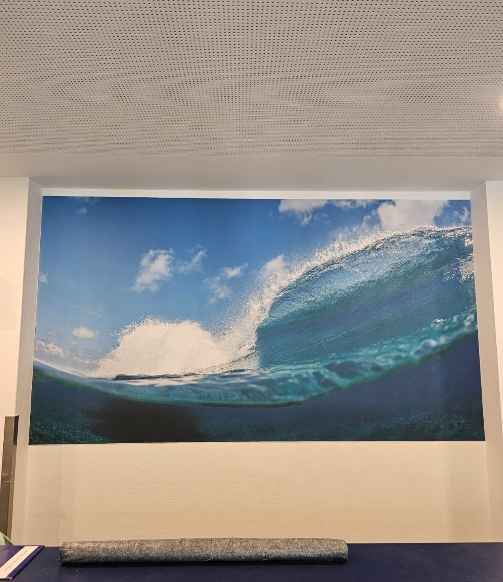Großes Wandbild einer brechenden Meereswelle – Welle rollt und spritzt Schaum; Wandbild hängt an weißer Innenwand unter perforierter Decke, Tisch mit aufgerolltem Stoff im Vordergrund.