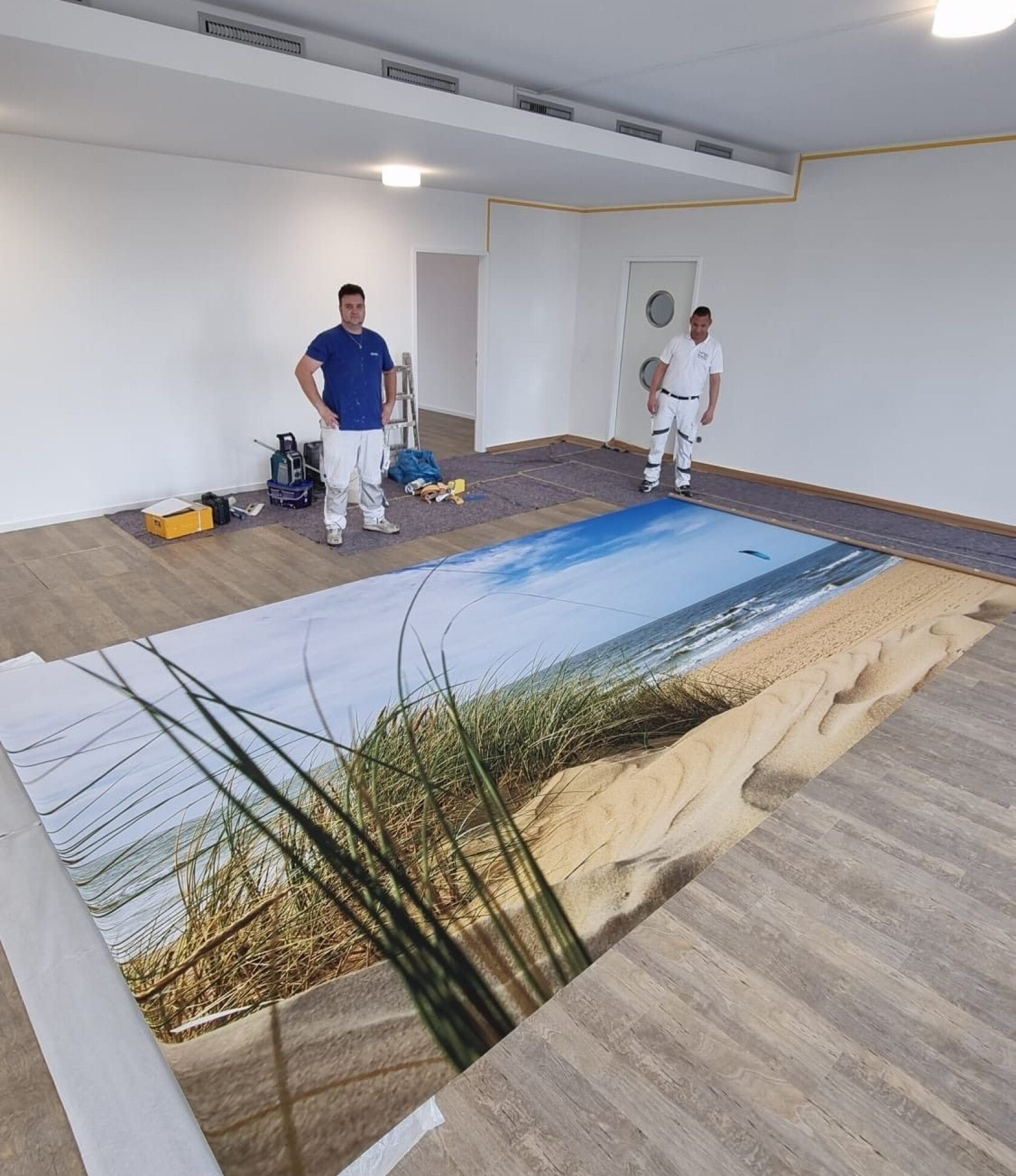 Großes Bodendruckmotiv einer Stranddüne wird auf Laminatboden ausgelegt — zwei Handwerker stehen daneben; leere weiße Räume, Werkzeug, Leiter und Deckenbeleuchtung sichtbar.
