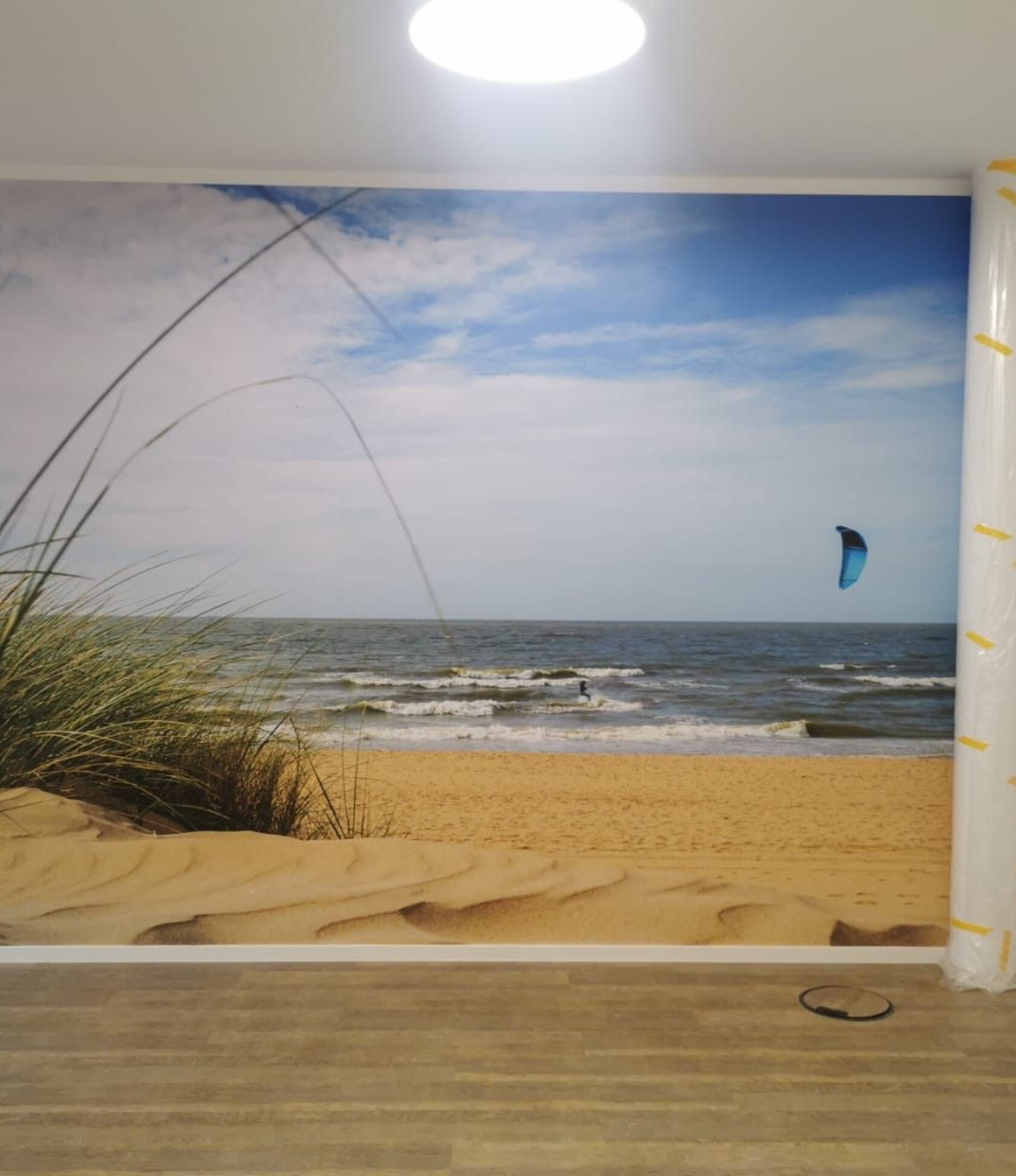 Große Wandfototapete zeigt Sanddünen und Strand mit Dünengras, Wellen und blauem Kitesegel; Raum mit Holzfußboden, Deckenleuchte oben und rechts gerollte Schutzfolie.