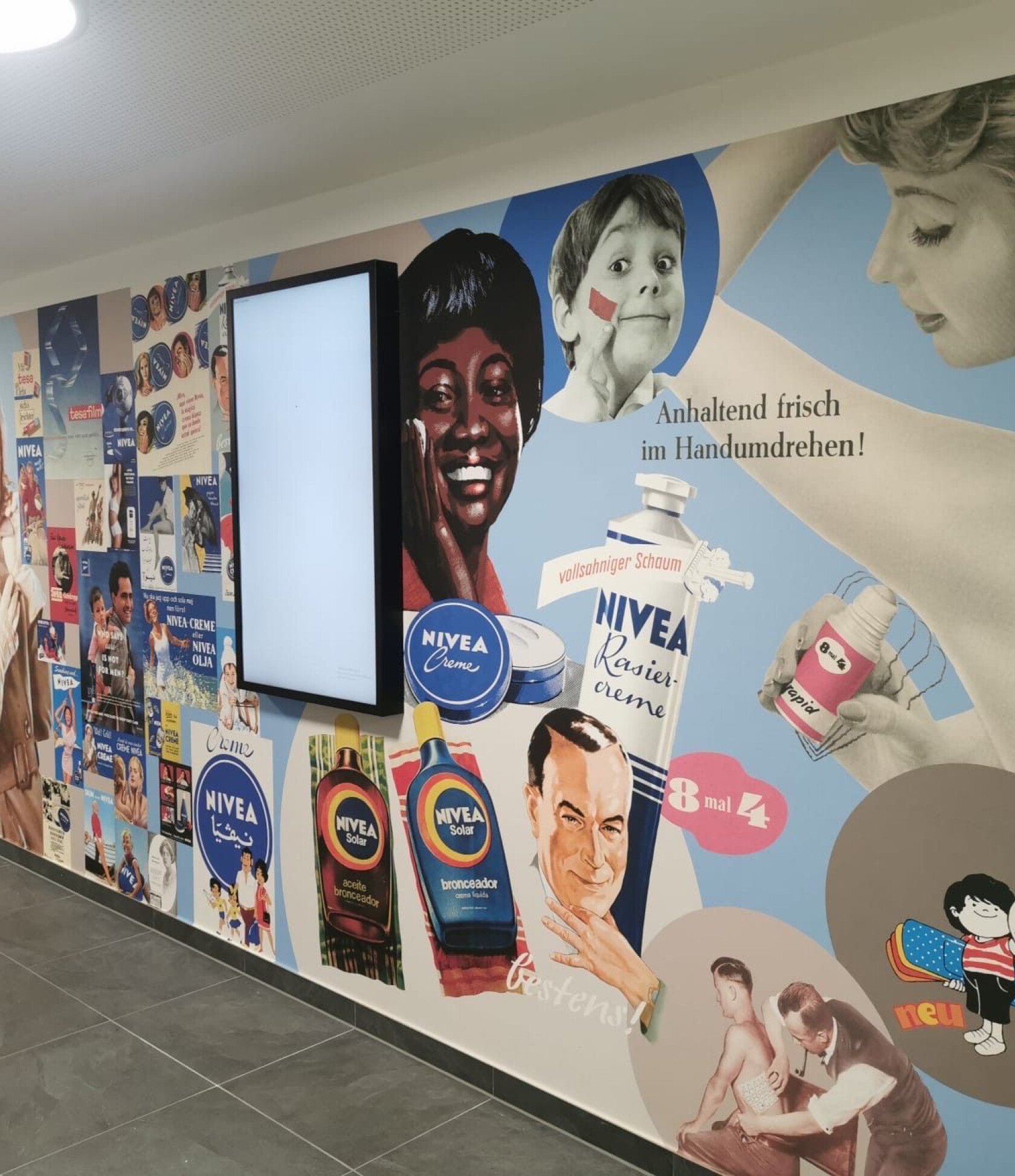 Wandcollage mit NIVEA-Werbemotiven – zeigt retro-Illustrationen von Gesichtern, Dosen und Flaschen neben einer leuchtenden Displaytafel in einem Innenflur; sichtbare Texte: 