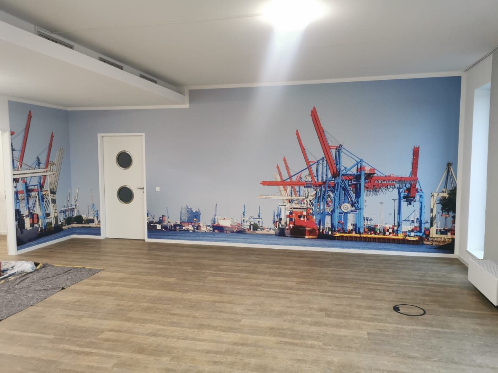 Große Wandfototapete eines Containerhafens – zeigt rote und blaue Hafenkräne über einem Frachtschiff; im Vordergrund leerer Raum mit hellem Holzfußboden, weiße Tür mit zwei Bullaugen.