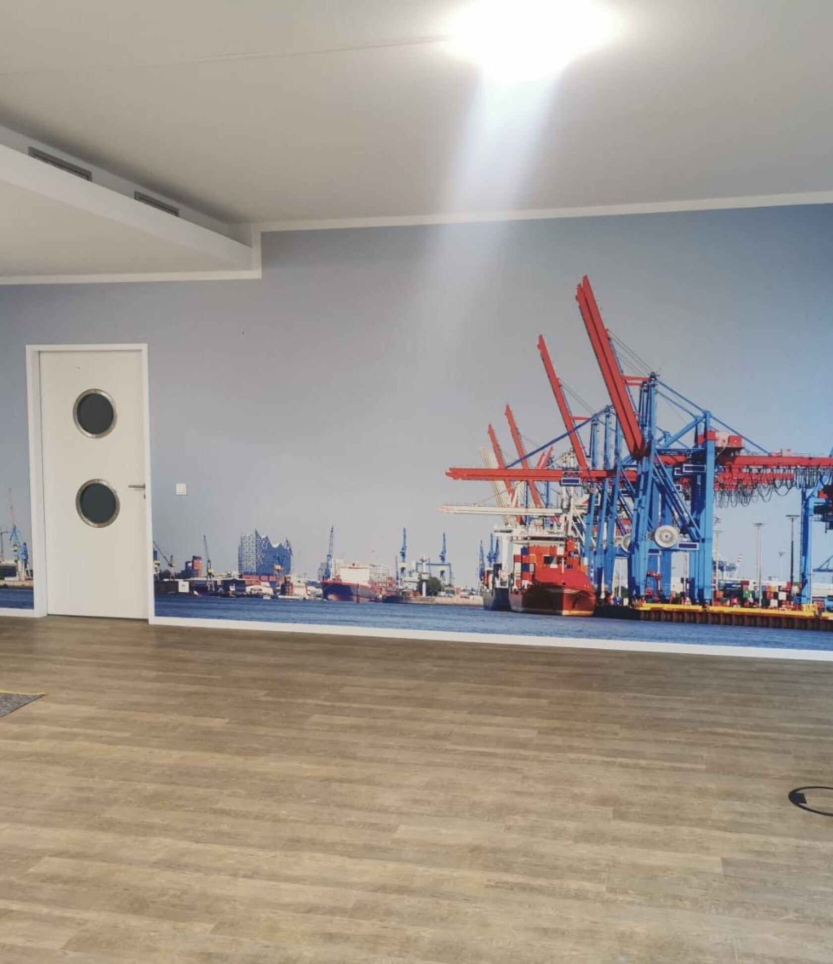 Große Wandfototapete eines Containerhafens – zeigt rote und blaue Hafenkräne über einem Frachtschiff; im Vordergrund leerer Raum mit hellem Holzfußboden, weiße Tür mit zwei Bullaugen.