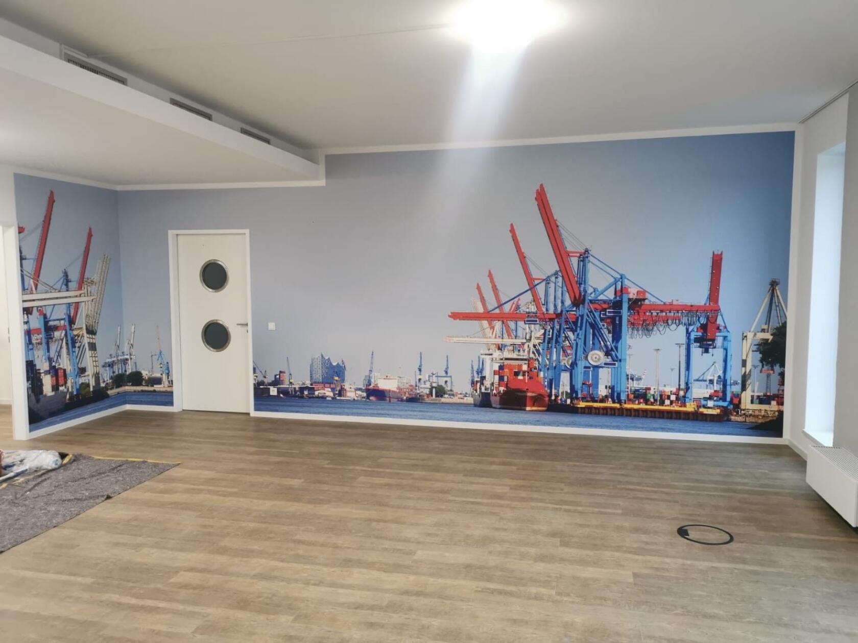 Große Wandfototapete eines Containerhafens – zeigt rote und blaue Hafenkräne über einem Frachtschiff; im Vordergrund leerer Raum mit hellem Holzfußboden, weiße Tür mit zwei Bullaugen.