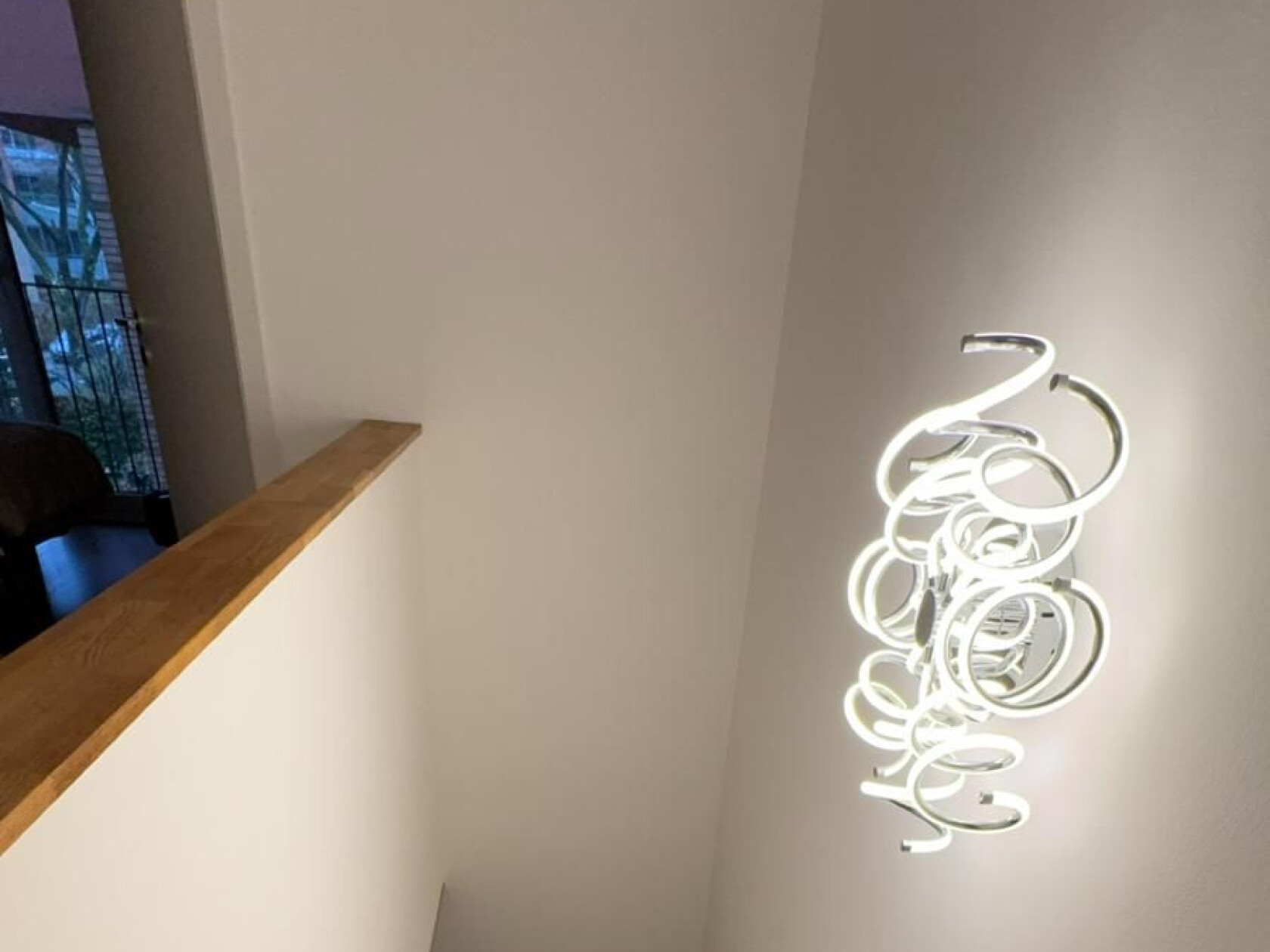 Moderne Deckenleuchte aus vielen beleuchteten weißen LED-Ringen, hängt an der Decke über einem Treppenabsatz; weiße Wände, sichtbares Holzgeländer und ein Fenster mit Balkon links.