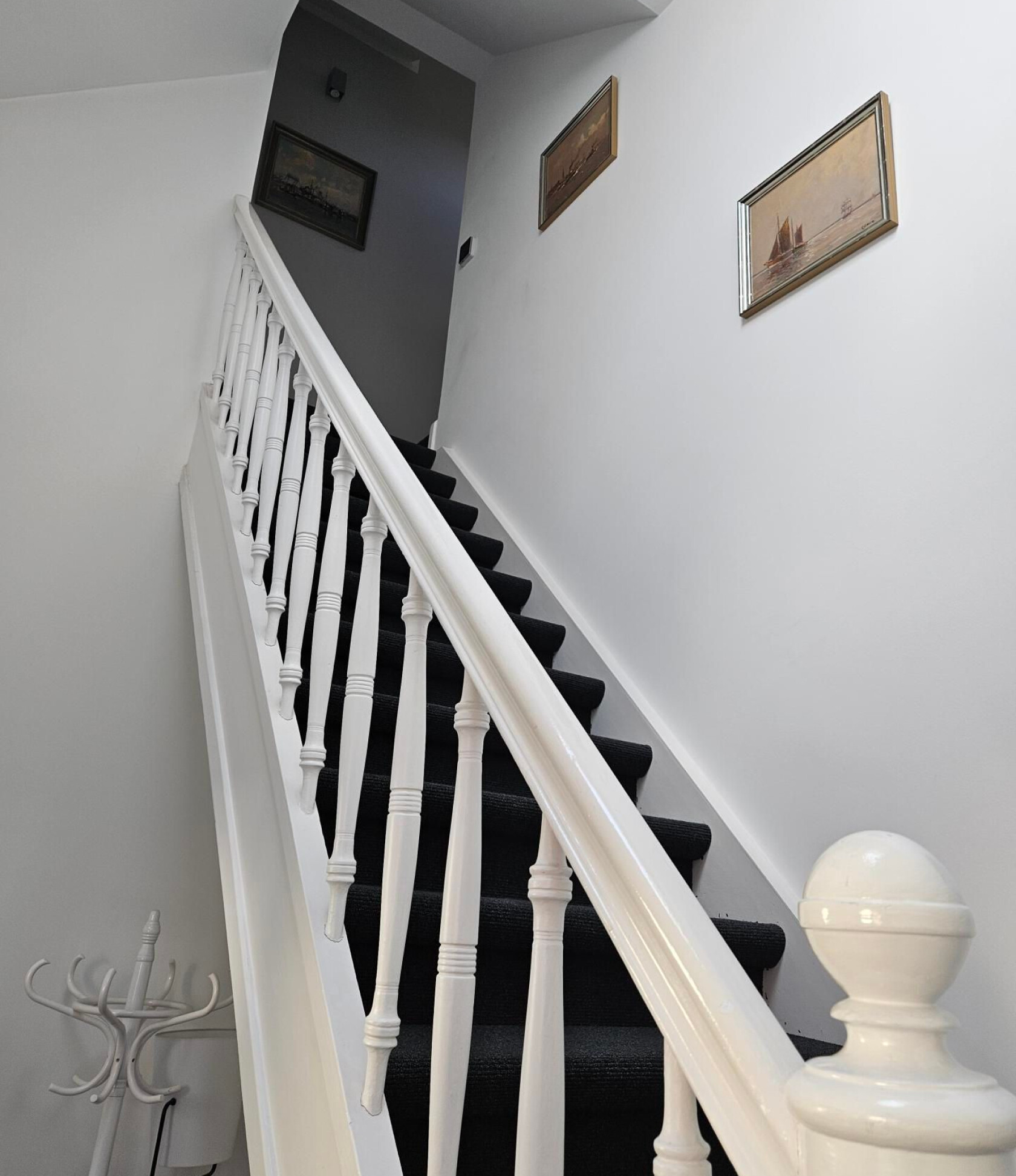 Treppe mit schwarzem Teppich, die nach oben zu einem dunklen Treppenabsatz führt; weiße Holzhandläufe und gedrehte Baluster, drei gerahmte Bilder mit Schiffsmotiven an der weißen Wand, Garderobenständer unten links.
