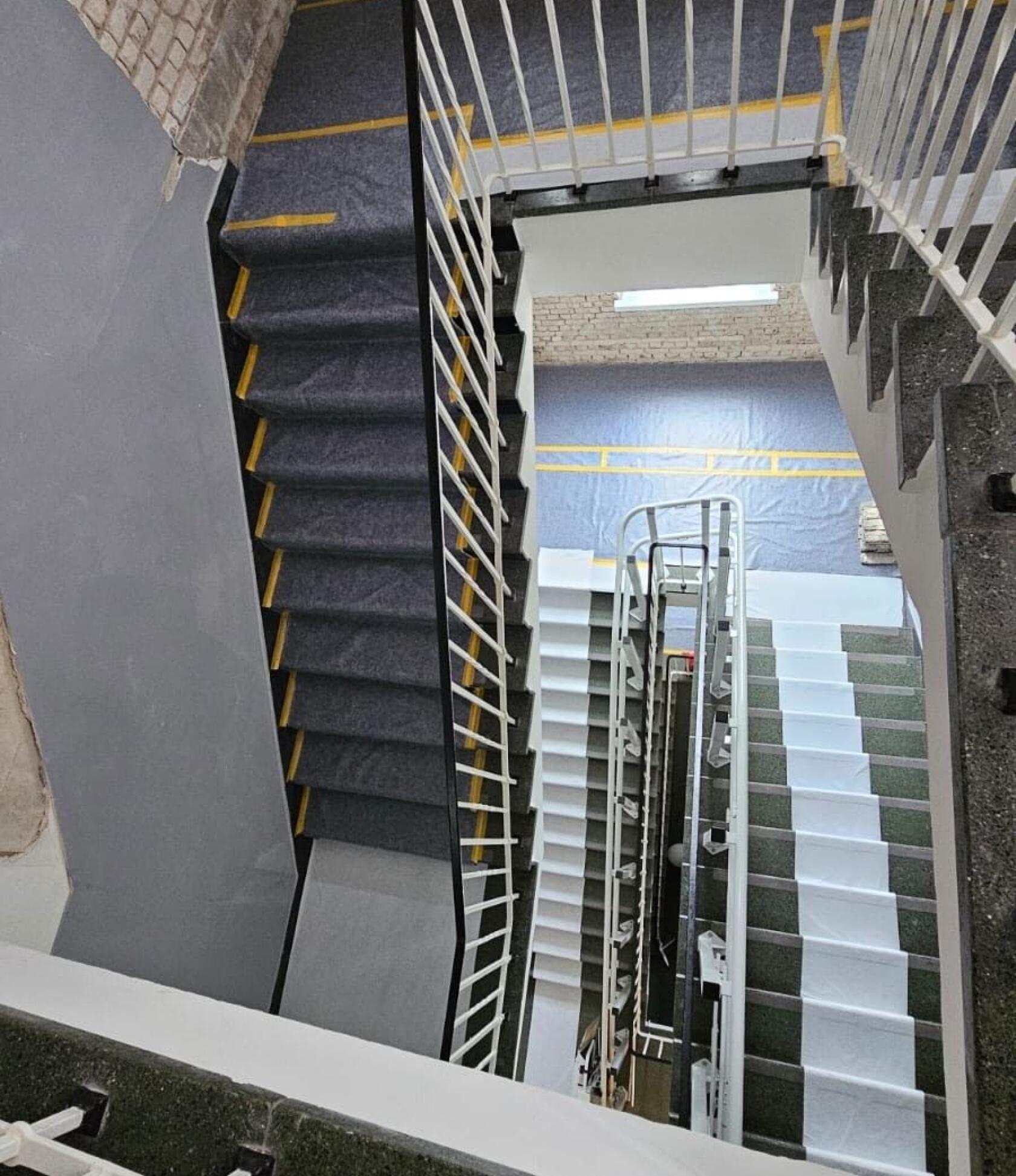 Mehrstöckiges Treppenhaus — Blick nach unten; Treppenabschnitte mit dunklem Teppich und weißen Schutzbahnen verlaufen spiralförmig; weiße Metallgeländer, Sicht auf Backsteinwand und gelbe Markierungsstreifen.