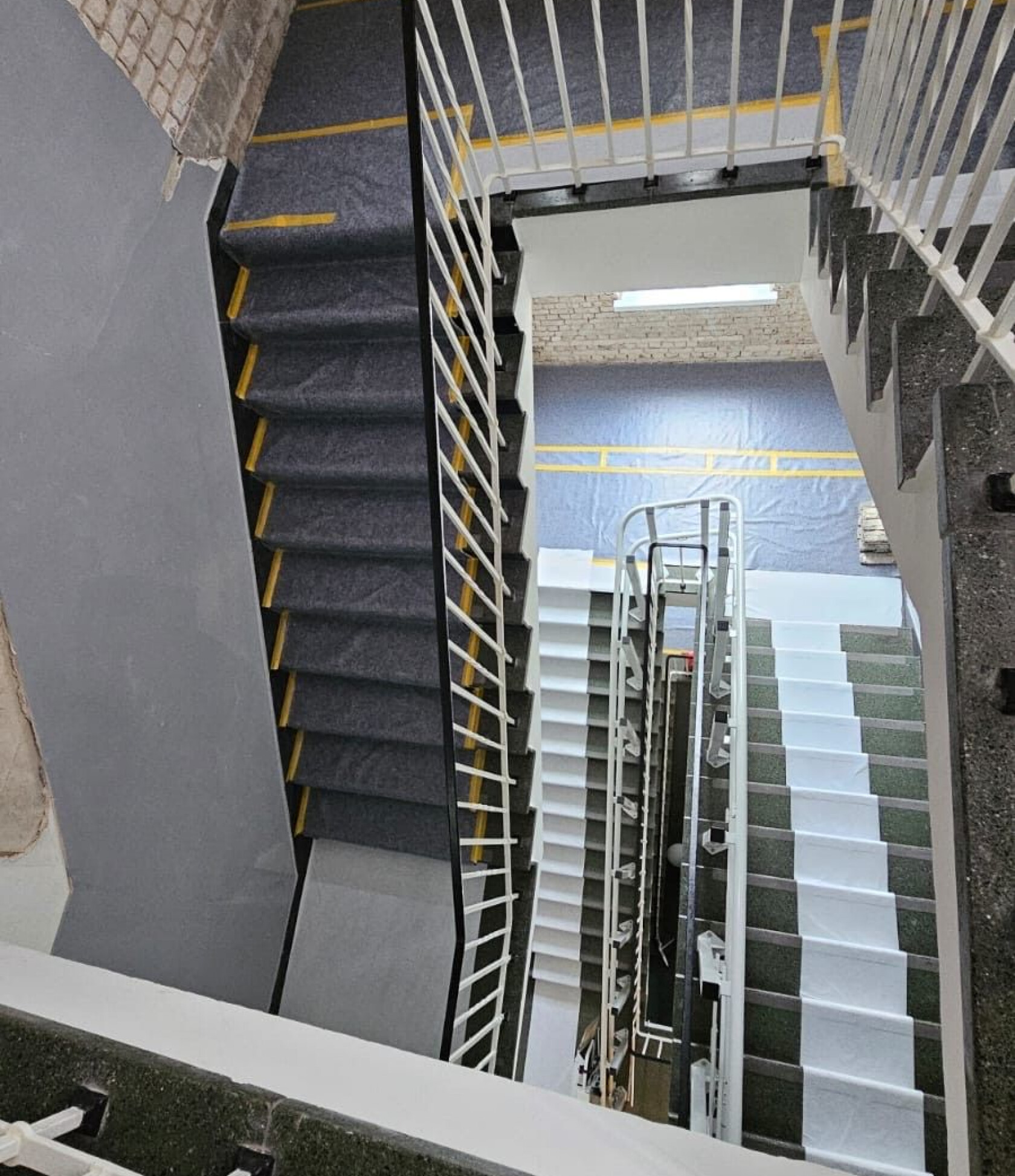 Mehrstöckiges Treppenhaus — Blick nach unten; Treppenabschnitte mit dunklem Teppich und weißen Schutzbahnen verlaufen spiralförmig; weiße Metallgeländer, Sicht auf Backsteinwand und gelbe Markierungsstreifen.