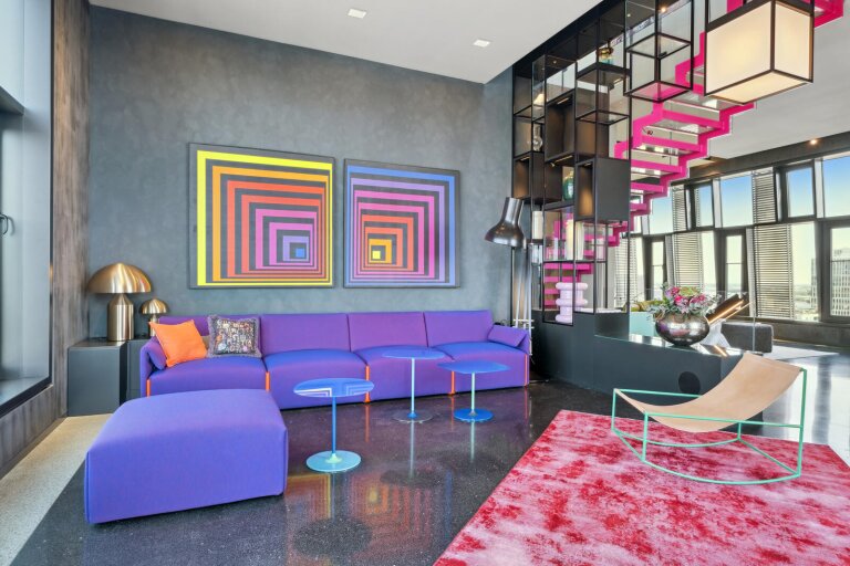 Violette modulare Couch mit Ottomane steht vor grauer Wand unter zwei bunten konzentrischen Quadratbildern; blaue Beistelltische, offenes Regal, pinke Treppe, roter Teppich vor großen Fenstern.