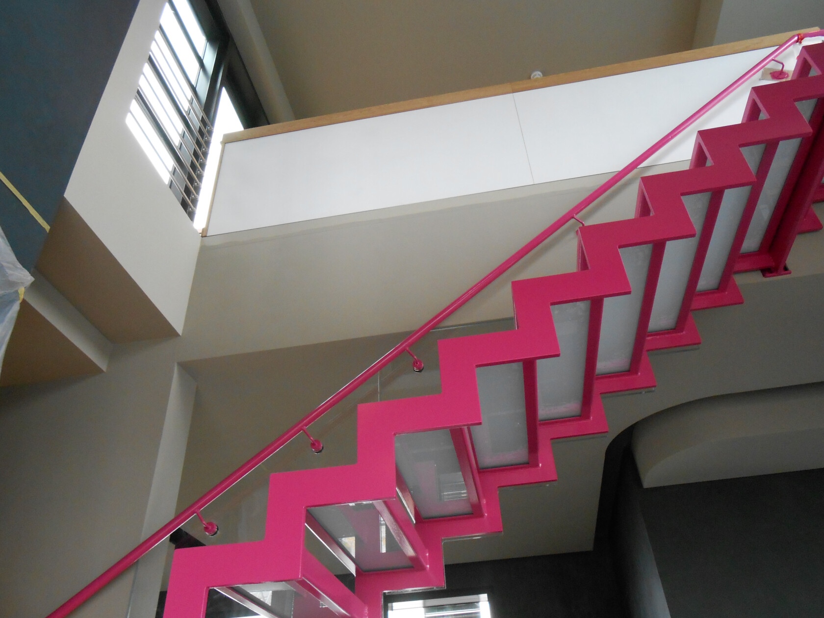 Pink lackierte, gezackte Stahltreppe verläuft diagonal nach oben; rechteckige Glasunterseiten der Stufen sichtbar, links hohes Fenster mit Lamellen, weiße Brüstung und graue Innenwände.
