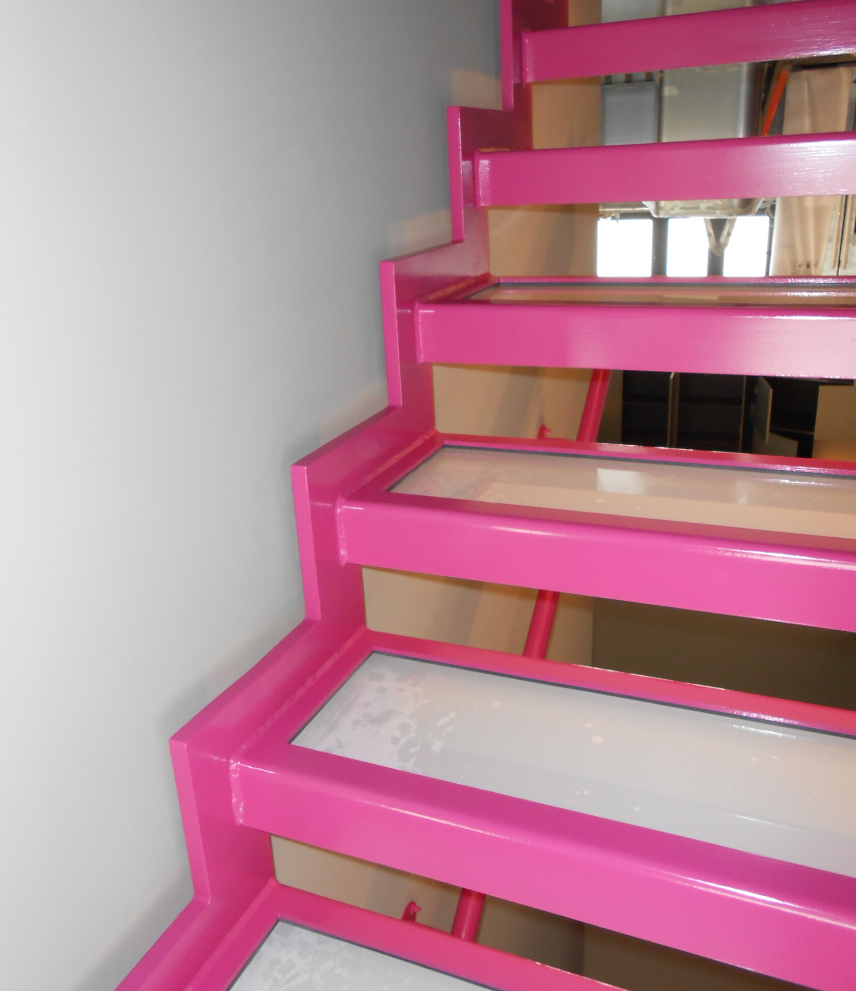 Pink lackierte Metalltreppe mit rechteckigen Glasstufen, verläuft diagonal nach oben; links glatte helle Wand, rechts Durchblick auf Fenster und technische Installationen des Treppenhauses.