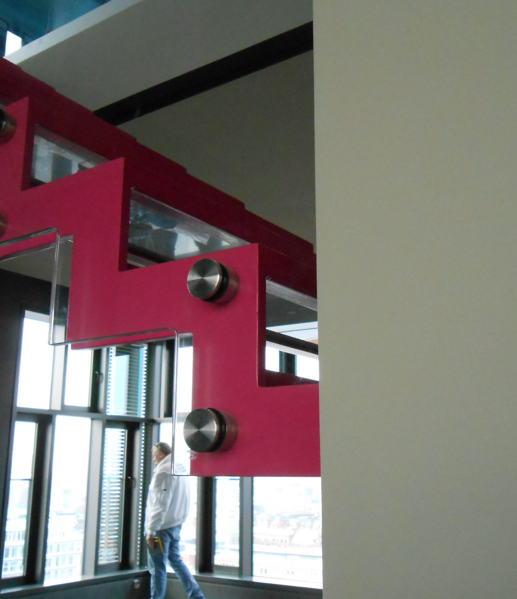 Zickzackförmiges rosa Metallträgerstück mit runden Edelstahlbefestigungen hält eingesetzte Glasscheiben — angebracht an Innenwand neben großen Fenstern, im Hintergrund stehende Person und Stadtblick.