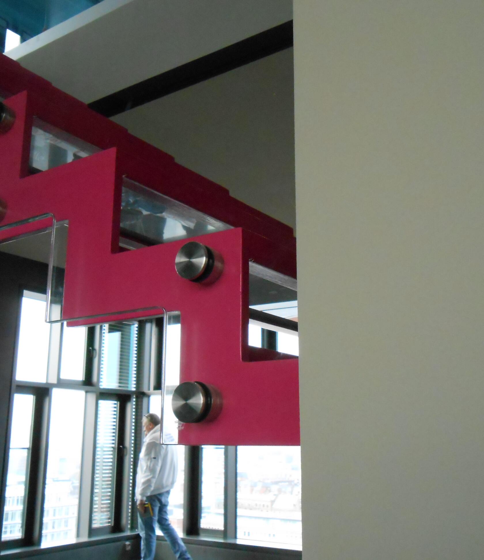 Zickzackförmiges rosa Metallträgerstück mit runden Edelstahlbefestigungen hält eingesetzte Glasscheiben — angebracht an Innenwand neben großen Fenstern, im Hintergrund stehende Person und Stadtblick.