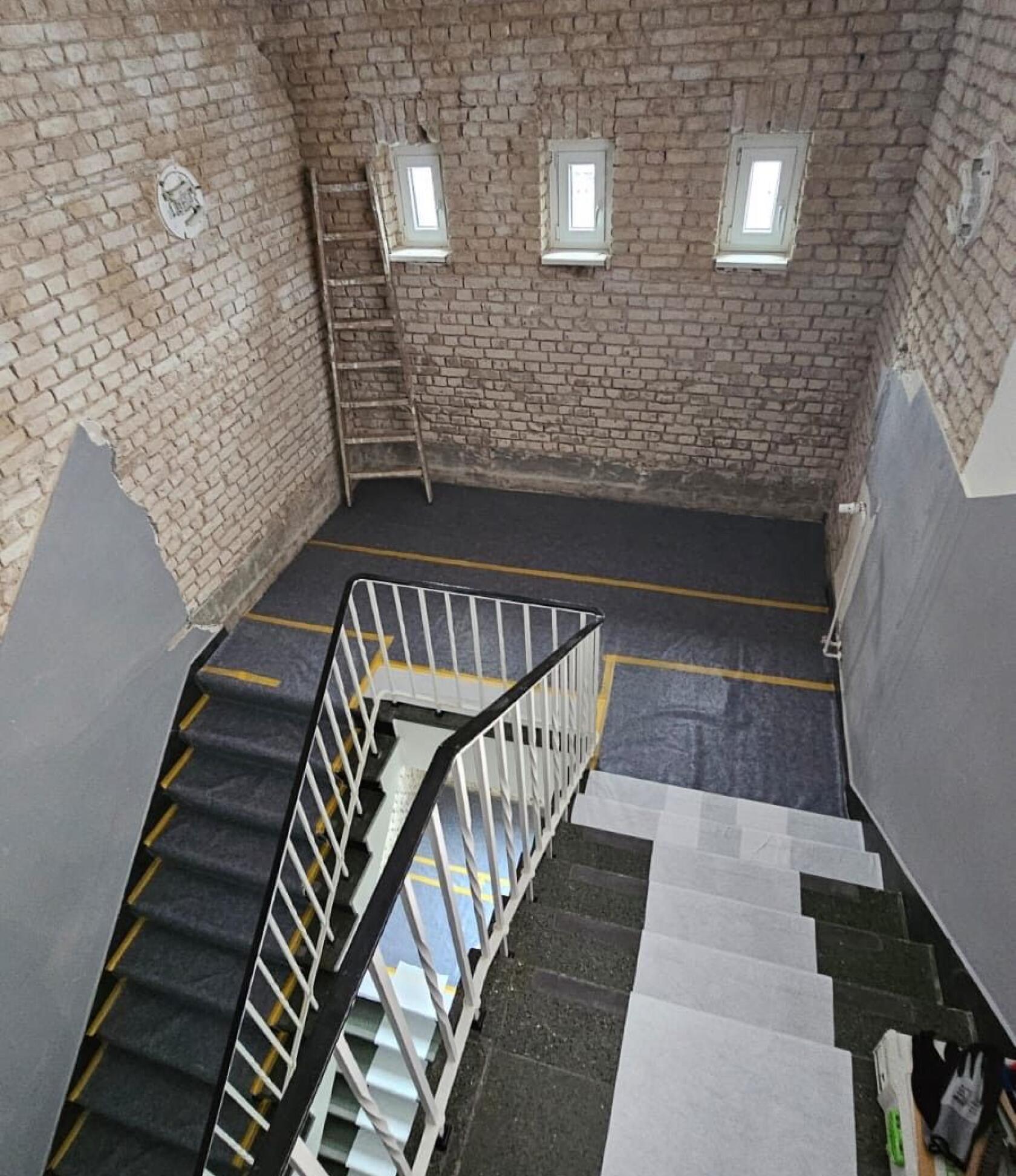 Geschwungene Treppe mit weißem Metallgeländer, nach unten führend; unverputzte Backsteinwände, drei kleine Fenster, eine Holzleiter an der Wand, grauer Bodenbelag mit gelben Markierungsstreifen und weißen Schutzfolien auf Stufen.