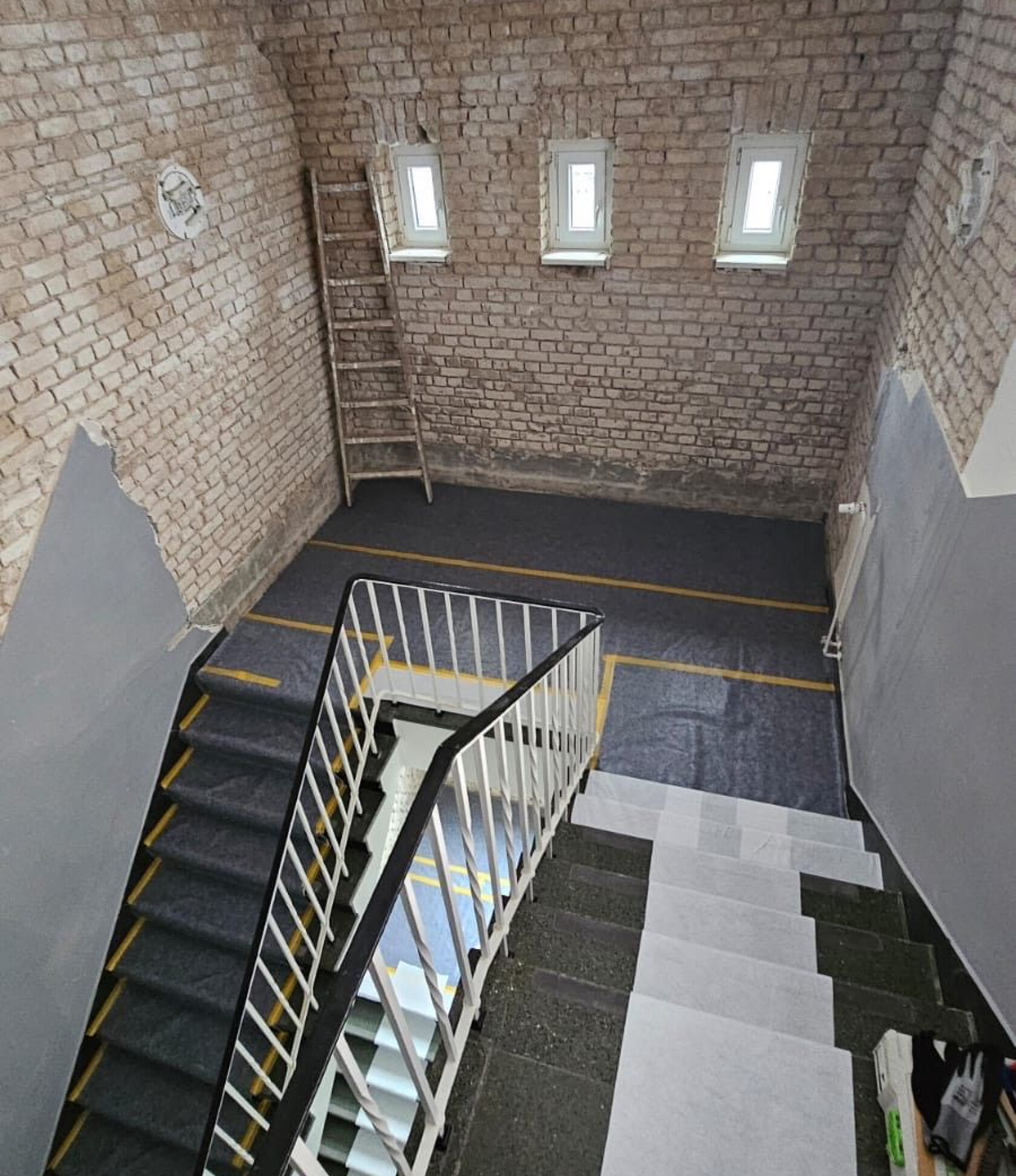 Geschwungene Treppe mit weißem Metallgeländer, nach unten führend; unverputzte Backsteinwände, drei kleine Fenster, eine Holzleiter an der Wand, grauer Bodenbelag mit gelben Markierungsstreifen und weißen Schutzfolien auf Stufen.