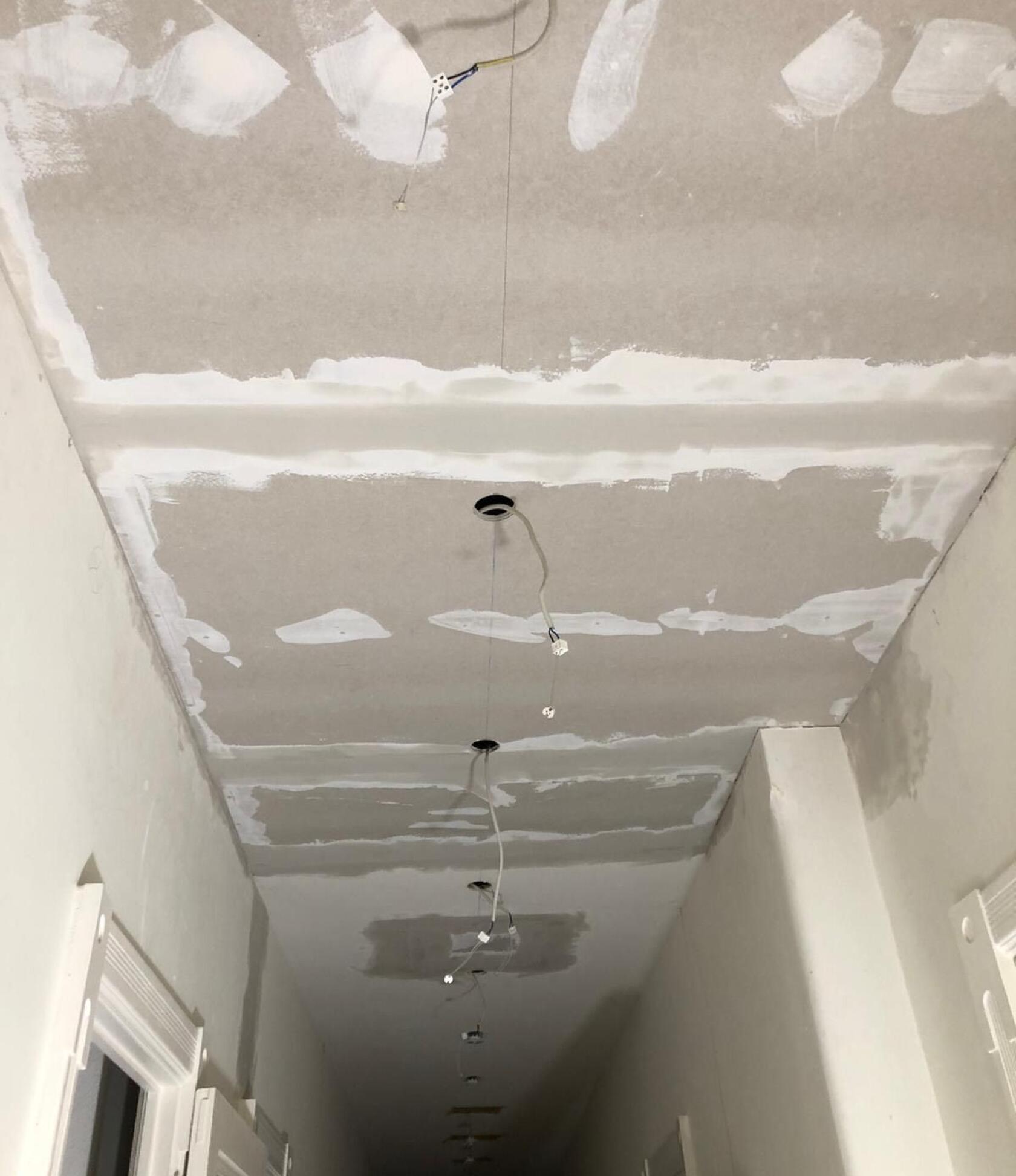 Decke aus Gipskartonplatten – gespachtelte Fugen und mehrere herabhängende Elektrokabel für Einbauleuchten – enger, schmaler Flur mit hellen Wänden und sichtbaren Türrahmen.