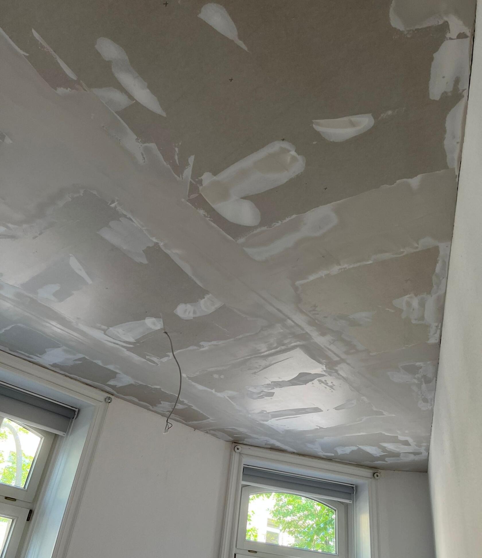 Decke mit verspachtelten Gipskartonfugen, frischen Spachtelstellen und sichtbaren Nahtkanten; loses Elektrokabel hängt herunter; zwei Fenster mit Rollläden und Tageslicht an angrenzender weißer Wand.