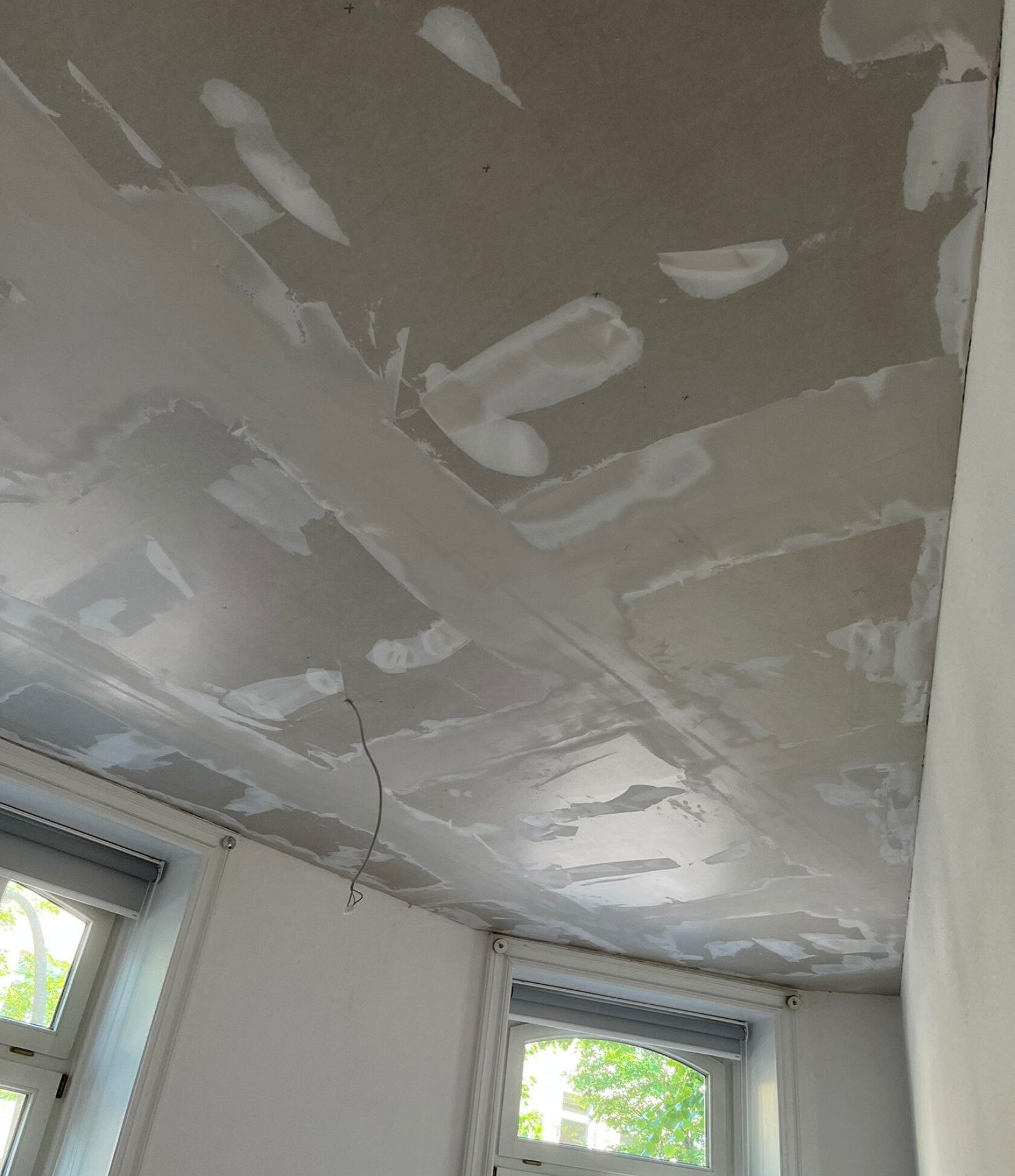 Decke mit verspachtelten Gipskartonfugen, frischen Spachtelstellen und sichtbaren Nahtkanten; loses Elektrokabel hängt herunter; zwei Fenster mit Rollläden und Tageslicht an angrenzender weißer Wand.