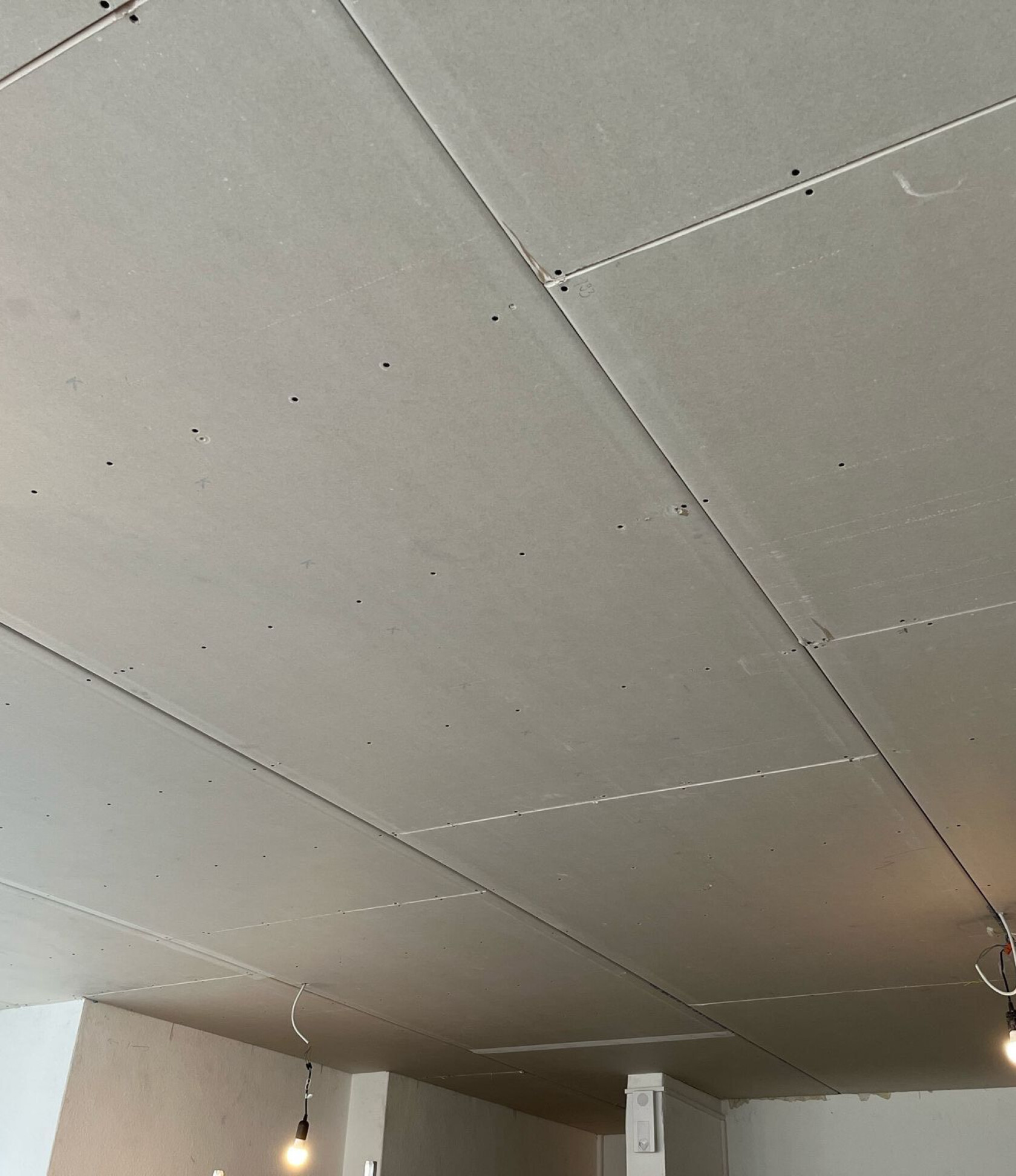 Gipskarton-Decke mit verschraubten Platten und sichtbaren Fugen, zwei hängende nackte Leuchtbirnen an freiliegenden Kabeln, Innenecke mit Fenster und teilweise verputzten Wänden.