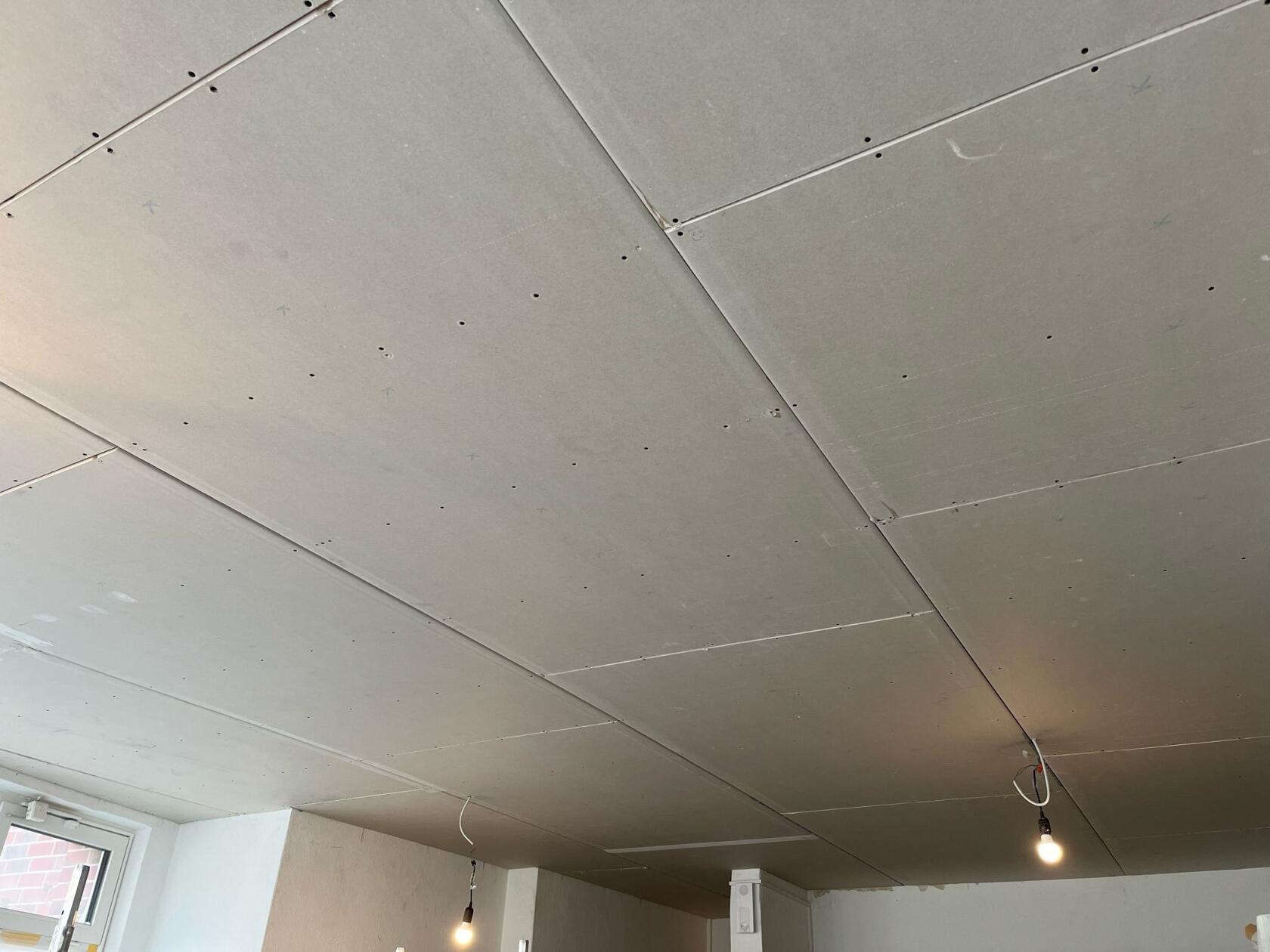 Gipskarton-Decke mit verschraubten Platten und sichtbaren Fugen, zwei hängende nackte Leuchtbirnen an freiliegenden Kabeln, Innenecke mit Fenster und teilweise verputzten Wänden.
