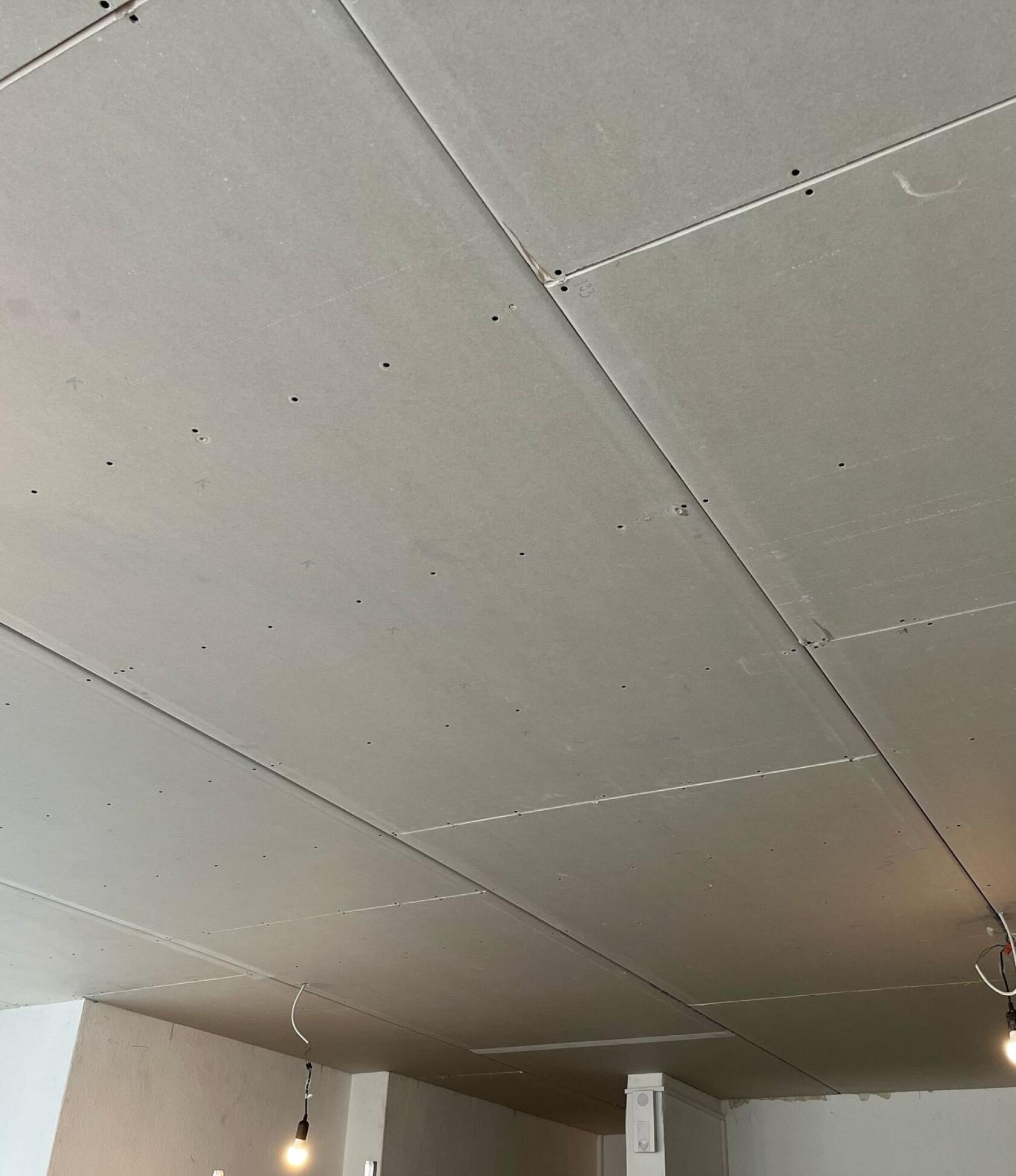Gipskarton-Decke mit verschraubten Platten und sichtbaren Fugen, zwei hängende nackte Leuchtbirnen an freiliegenden Kabeln, Innenecke mit Fenster und teilweise verputzten Wänden.
