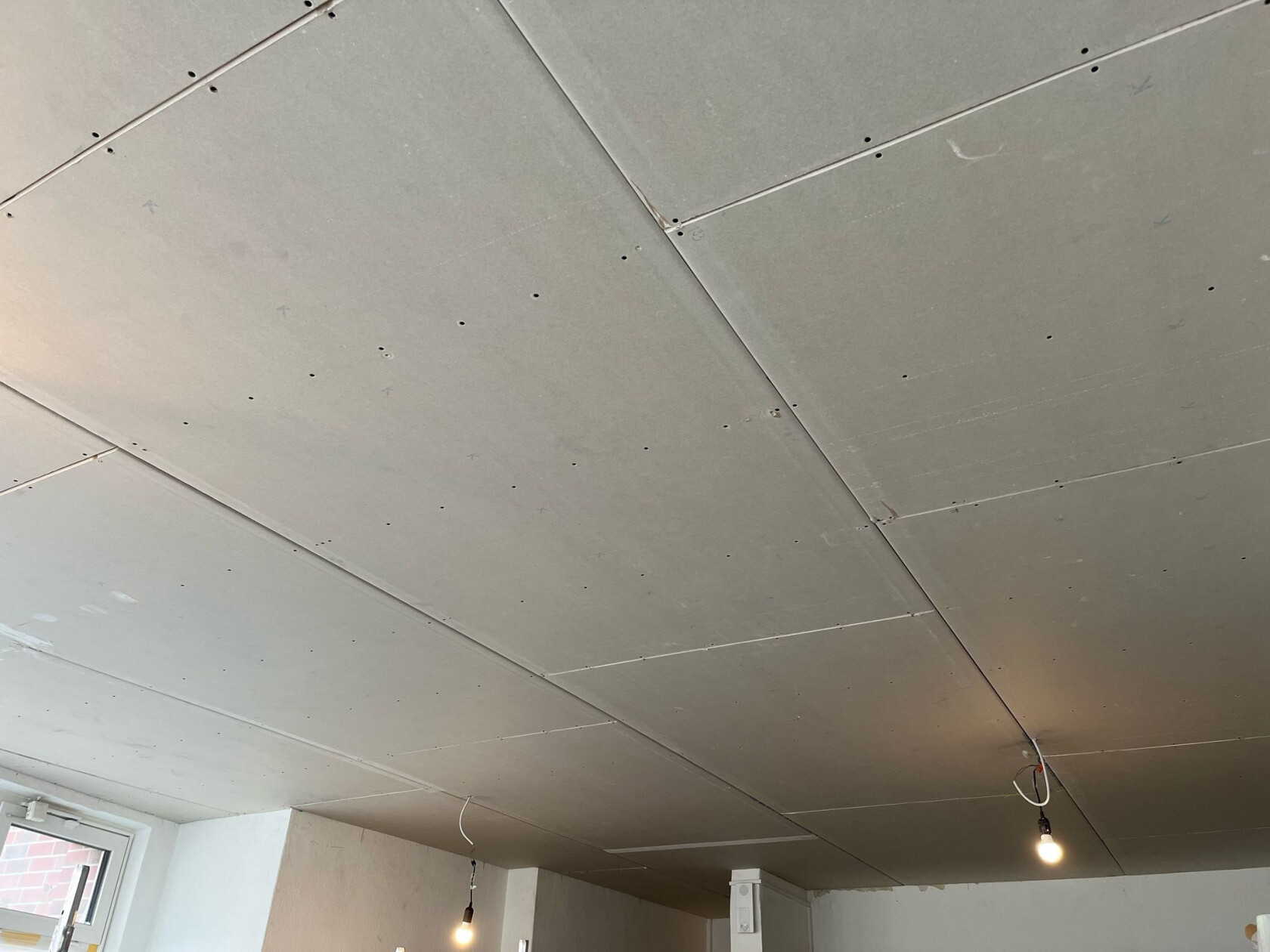 Gipskarton-Decke mit verschraubten Platten und sichtbaren Fugen, zwei hängende nackte Leuchtbirnen an freiliegenden Kabeln, Innenecke mit Fenster und teilweise verputzten Wänden.
