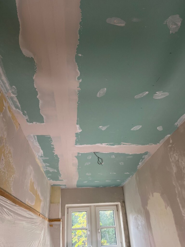 Grüne Gipskarton-Decke mit weiß gespachtelten Längs- und Querfugen, hängende Elektrokabel sichtbar; Innenraum unter Renovierung, unvollständig verputzte Wände, Fenster am Raumende.