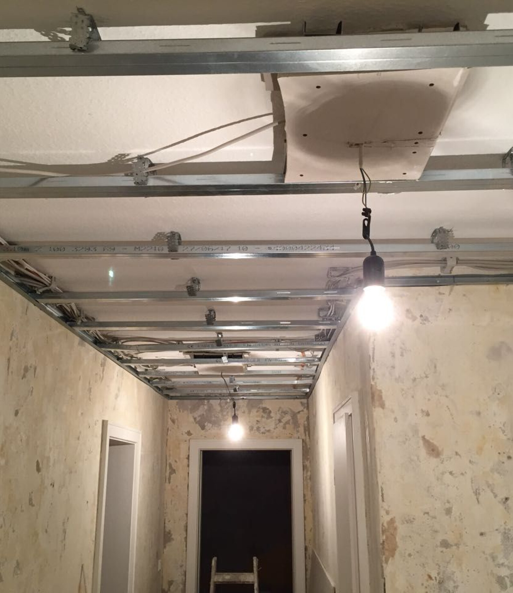 Metall-Unterkonstruktion für abgehängte Decke mit zwei hängenden Glühbirnen, eine leuchtet; schmaler Flur mit abgeblätterten Wänden, Türrahmen und Leiter am Ende.