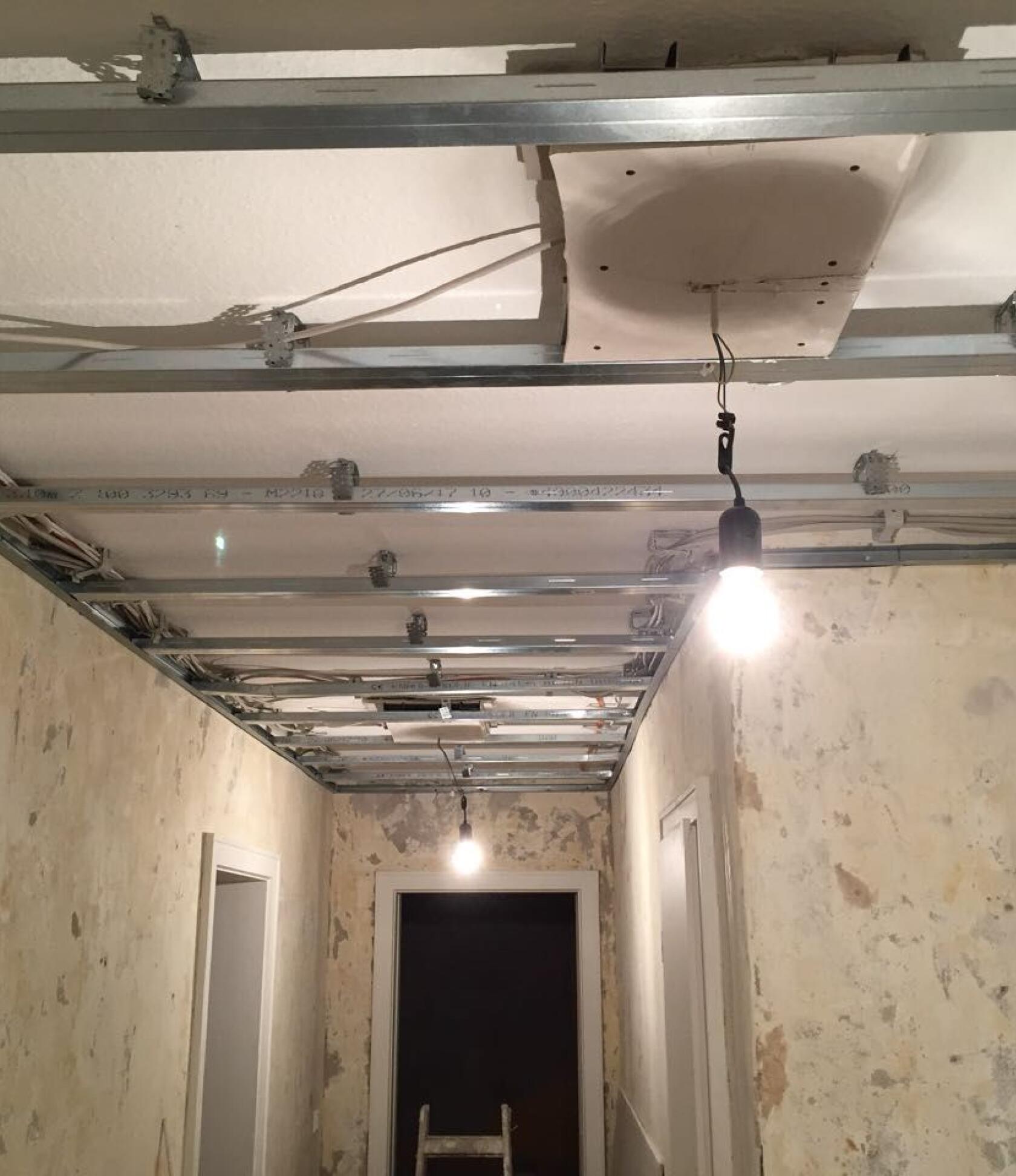 Metall-Unterkonstruktion für abgehängte Decke mit zwei hängenden Glühbirnen, eine leuchtet; schmaler Flur mit abgeblätterten Wänden, Türrahmen und Leiter am Ende.