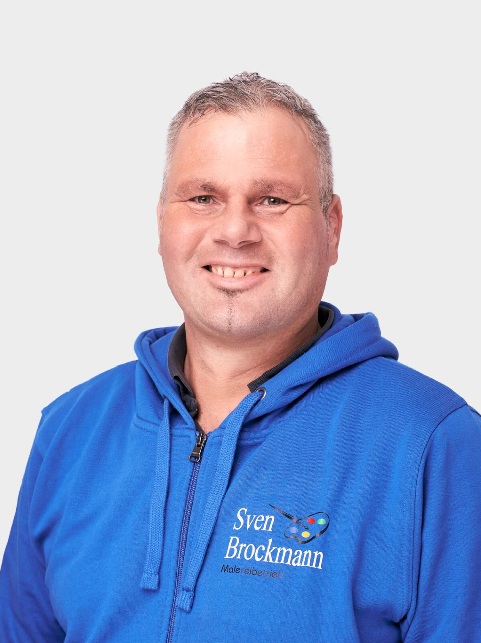 Mann mit kurzem grauem Haar, frontal zur Kamera stehend und leicht lächelnd; trägt blaue Kapuzenjacke mit Schrift 