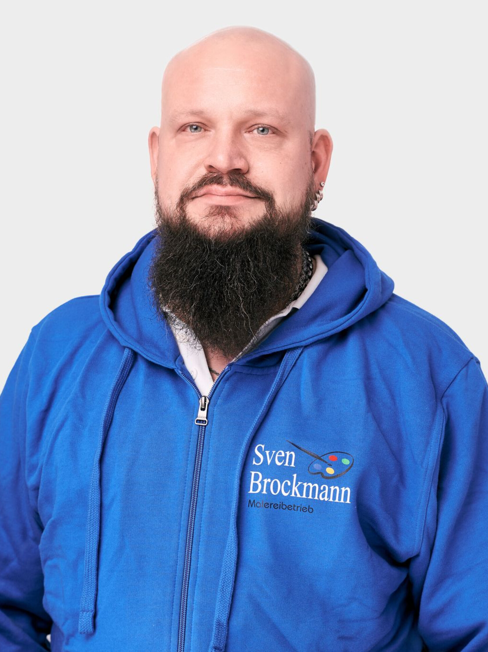 Bärtige, glatzköpfige Person mit langem dunklen Bart und mehreren Ohrringen schaut frontal; trägt blaue Kapuzenjacke mit gesticktem Schriftzug 