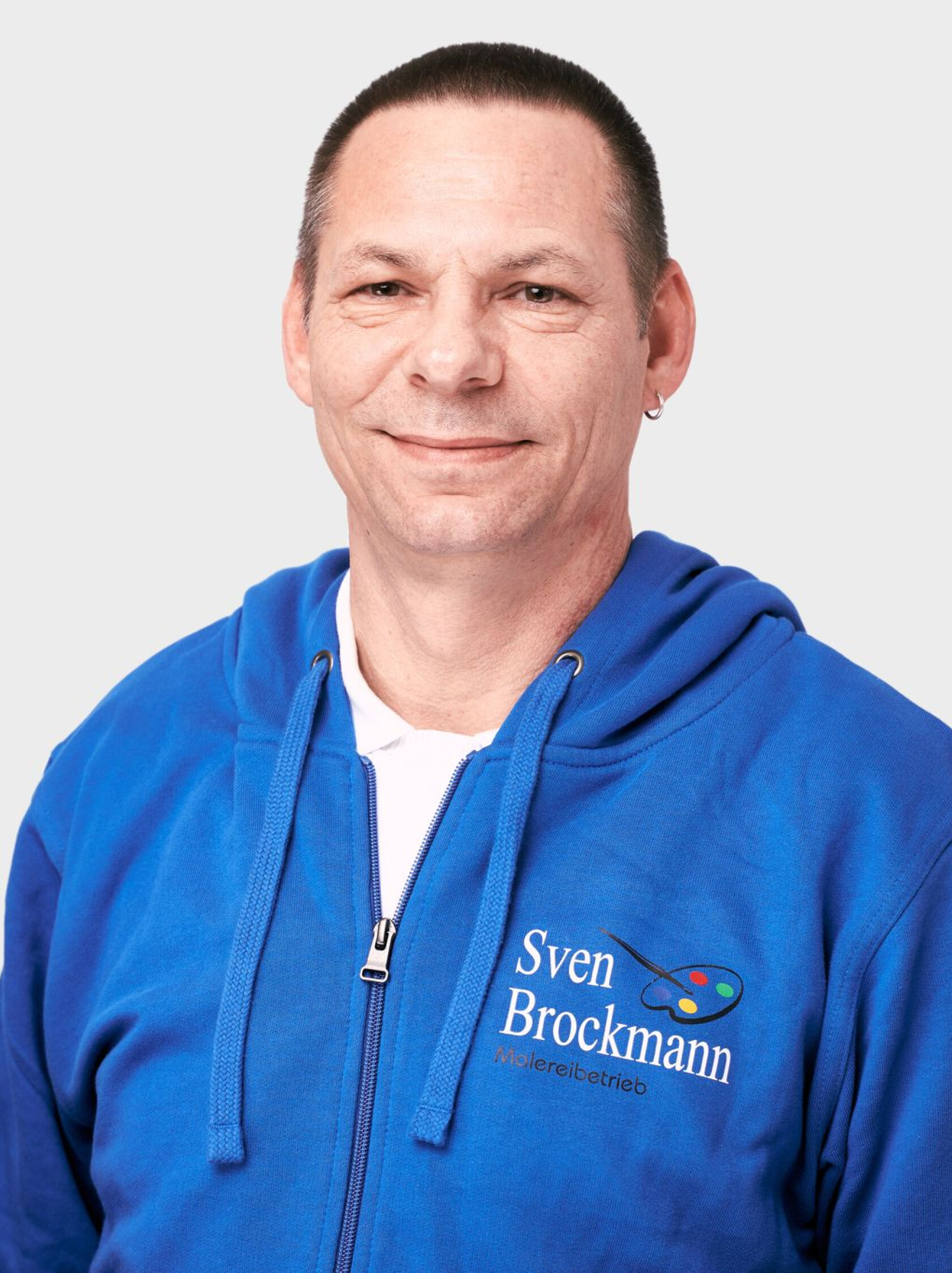 Mann blickt in die Kamera, trägt blaues Reißverschluss-Hoodie mit Malerpalette-Logo und weißem T-Shirt sichtbar vor hellem Hintergrund. Text auf Kleidung: 