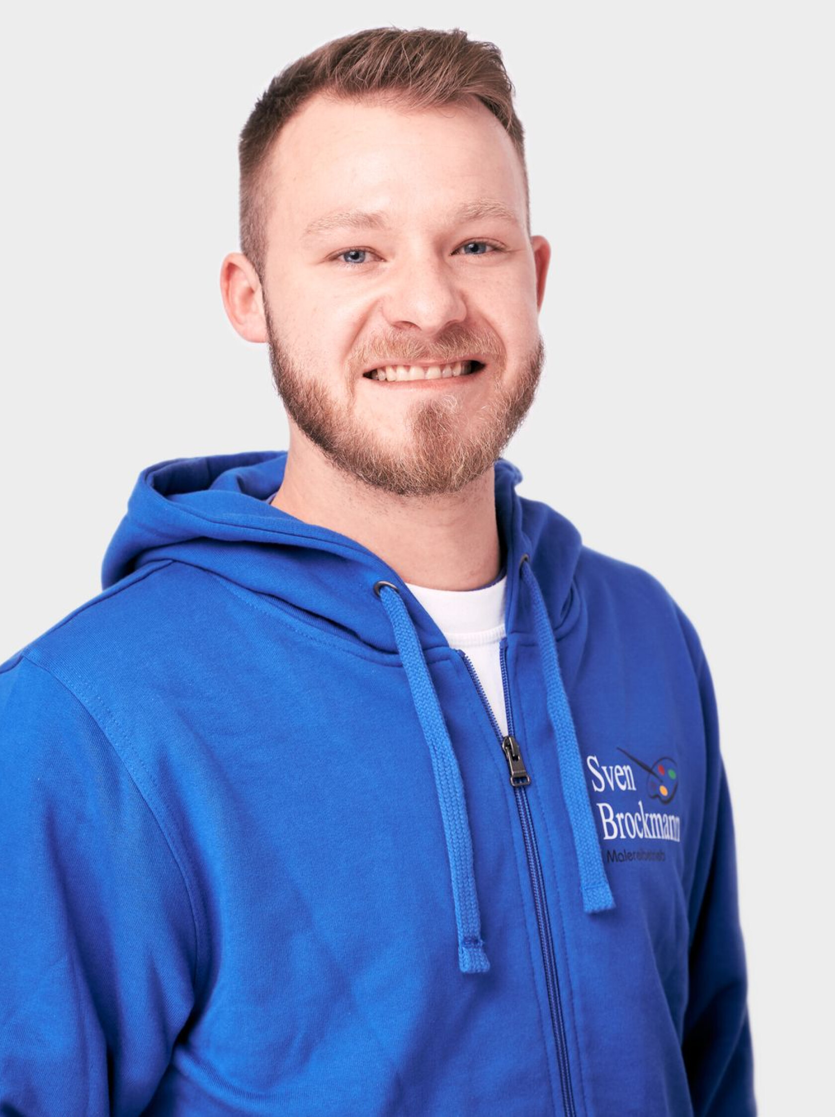 Junger Mann mit kurzem Haar und Bart – blickt zur Kamera und lächelt leicht – trägt blaues Kapuzen-Sweatshirt mit Reißverschluss und Logo 