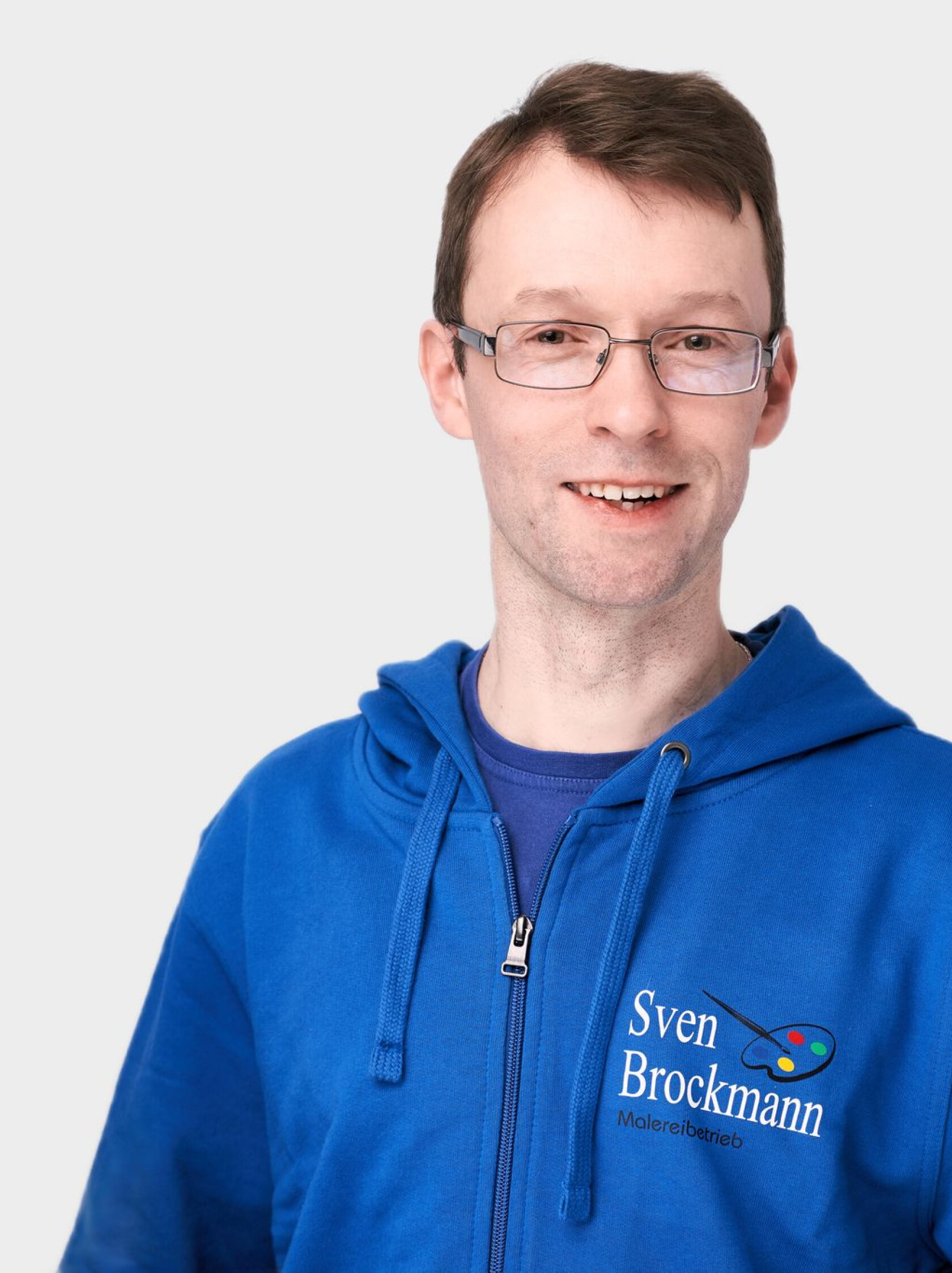 Mann mit Brille lächelt in die Kamera; trägt blaue Kapuzenjacke mit Reißverschluss, aufgedrucktem Text 'Sven Brockmann Malereibetrieb' und Farbpalette-Logo, vor hellem Hintergrund.