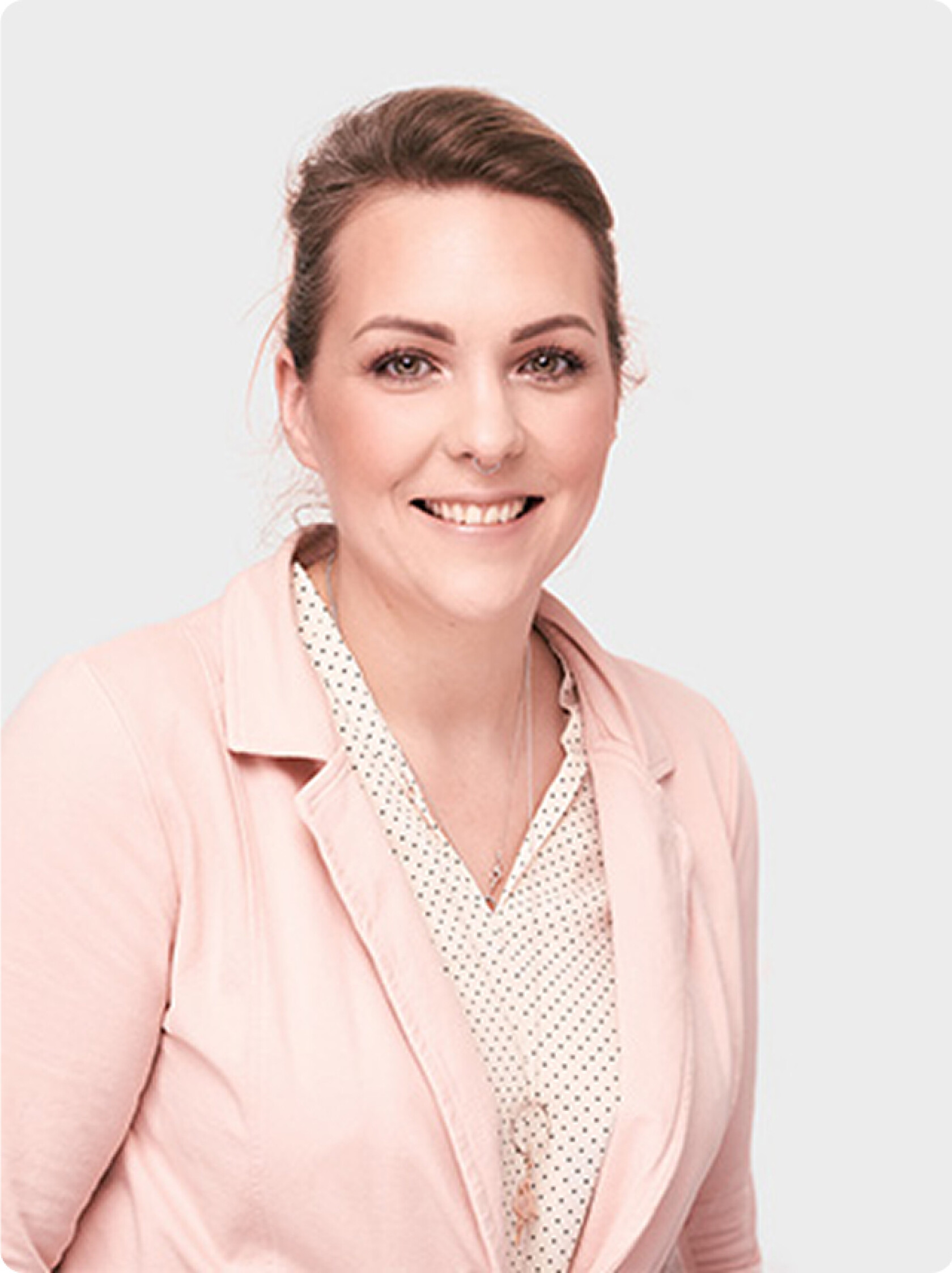 Frau, lächelnd, halbnahes Porträt vor hellgrauem Hintergrund; trägt hellrosa Blazer, weiße Bluse mit schwarzen Punkten, zurückgesteckte braune Haare und feine Silberkette.