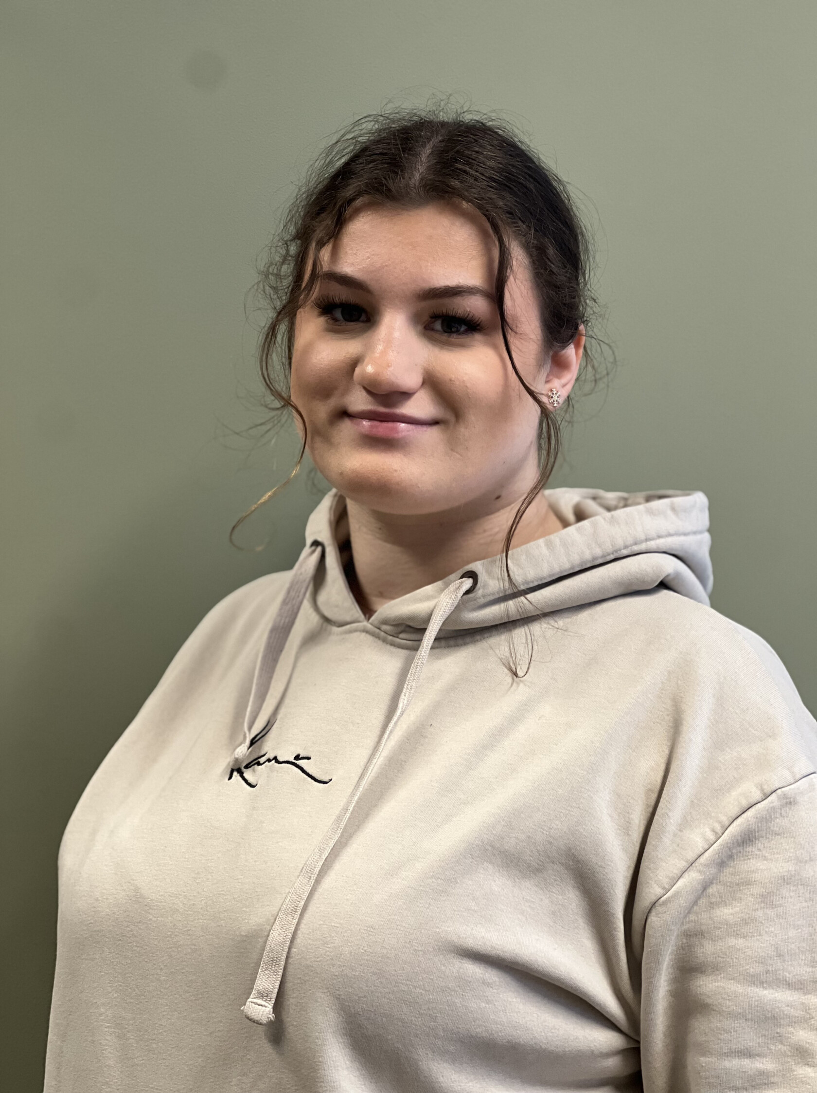 Frau blickt leicht zur Kamera, trägt beigen Kapuzenpullover mit Schriftzug 'Kane', dunkle Haare zu lockerem Knoten gebunden, mit kleinen Ohrsteckern sichtbar, vor einfarbiger grüner Wand.