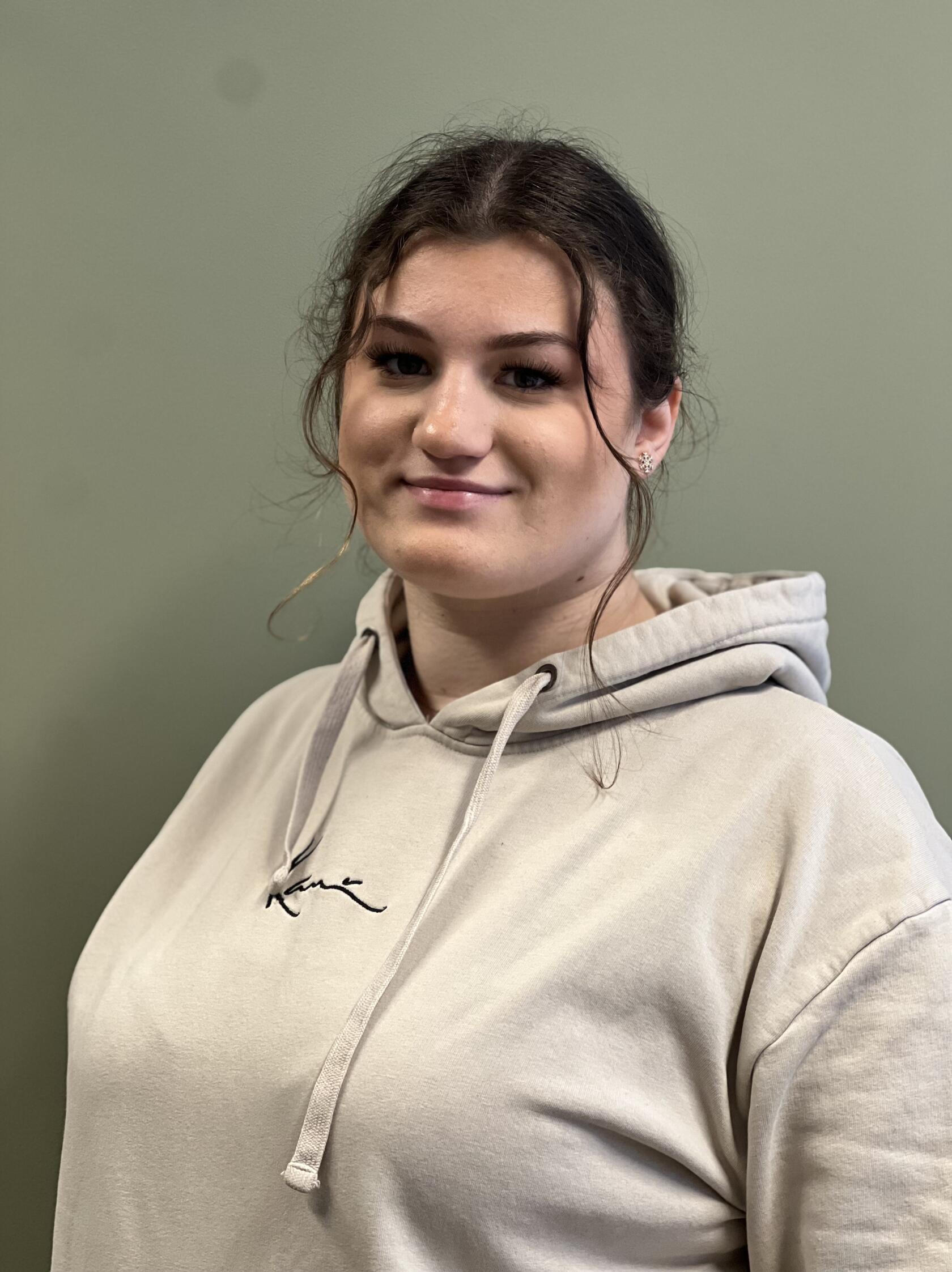 Frau blickt leicht zur Kamera, trägt beigen Kapuzenpullover mit Schriftzug 'Kane', dunkle Haare zu lockerem Knoten gebunden, mit kleinen Ohrsteckern sichtbar, vor einfarbiger grüner Wand.