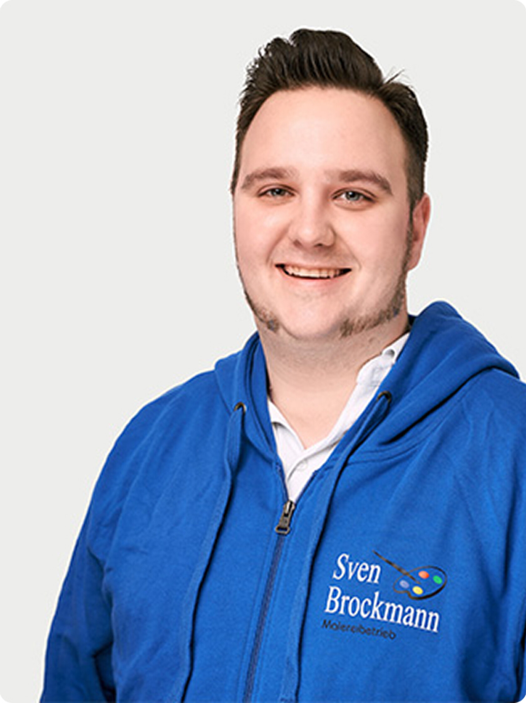 Mann, lächelt in die Kamera, halbkörpernah vor hellem einfarbigem Hintergrund; trägt blaues Kapuzen-Sweatshirt über weißem Hemd mit aufgesticktem Logo und Text 