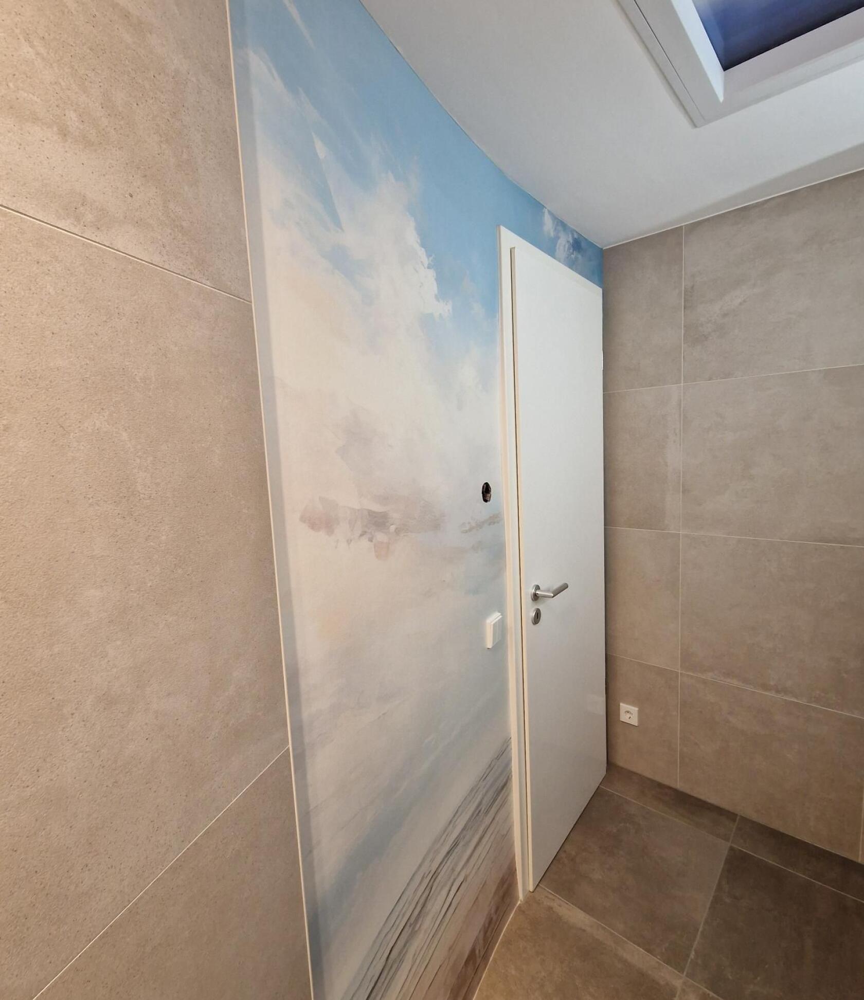Weiße Innentür mit Edelstahlgriff, eingebaut in Wand neben großformatigem Wandbild mit Himmel- und Meeresmotiv; Raum mit großen grauen Wand- und Bodenfliesen, Oberlicht.