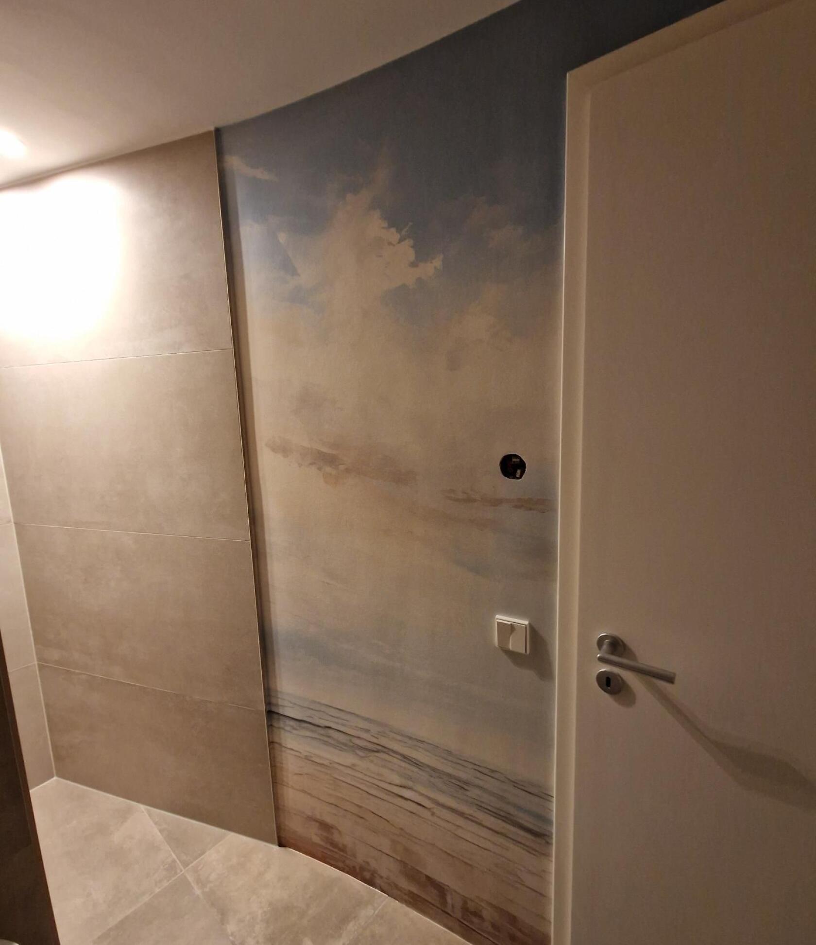 Wand mit großflächigem Himmel- und Küstenmotiv, das Wolken und horizontale Linien darstellt; rechts weiße Tür mit Griff, runde Öffnung, Lichtschalter, Fliesenboden.