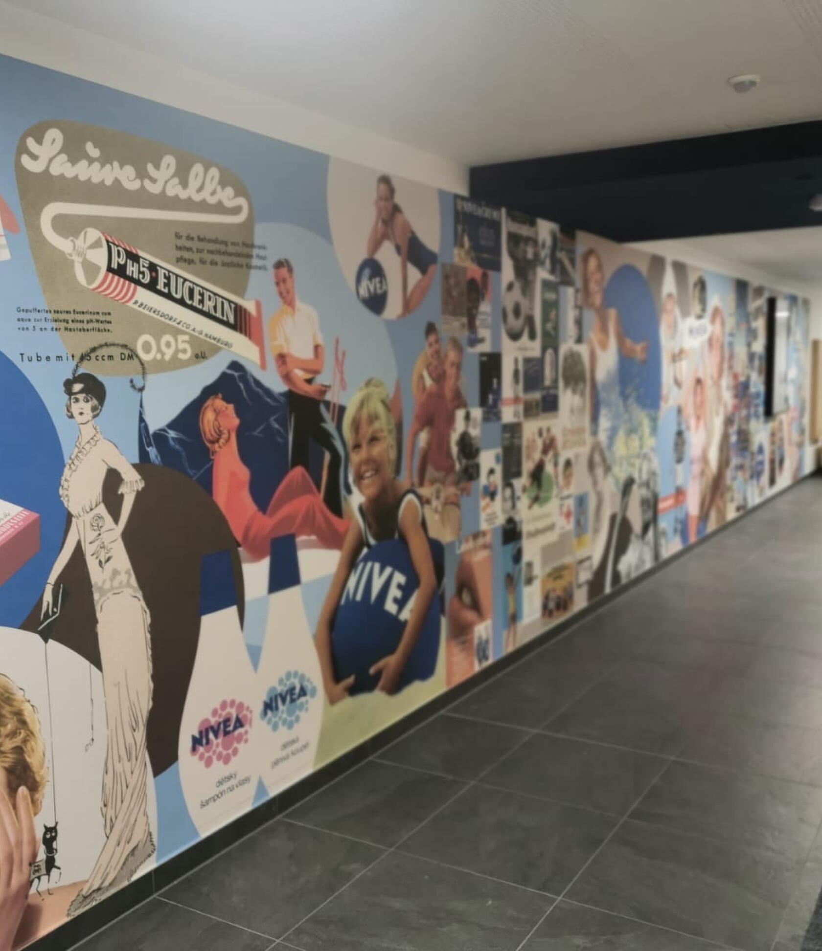 Bunte Wandcollage mit Retro-Werbemotiven und Personen – zeigt Nivea‑ und Eucerin‑Anzeigen auf einer langen Flurwand – heller Korridor mit dunkler Decke und grauem Fliesenboden. Text im Bild: 